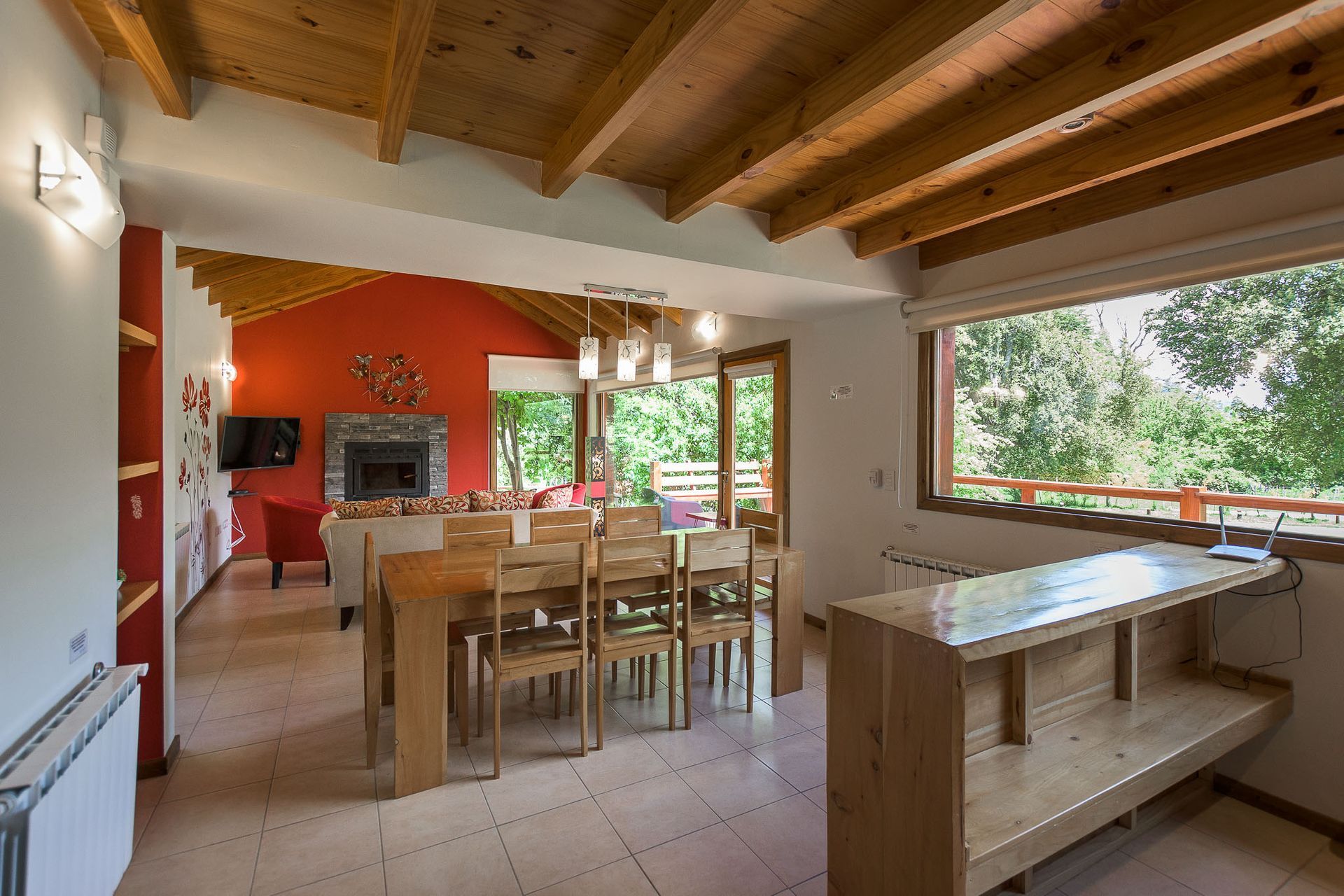 Sala de estar y comedor de planta abierta con techo rústico de vigas de madera, mesa de comedor de madera, chimenea y grandes ventanales.