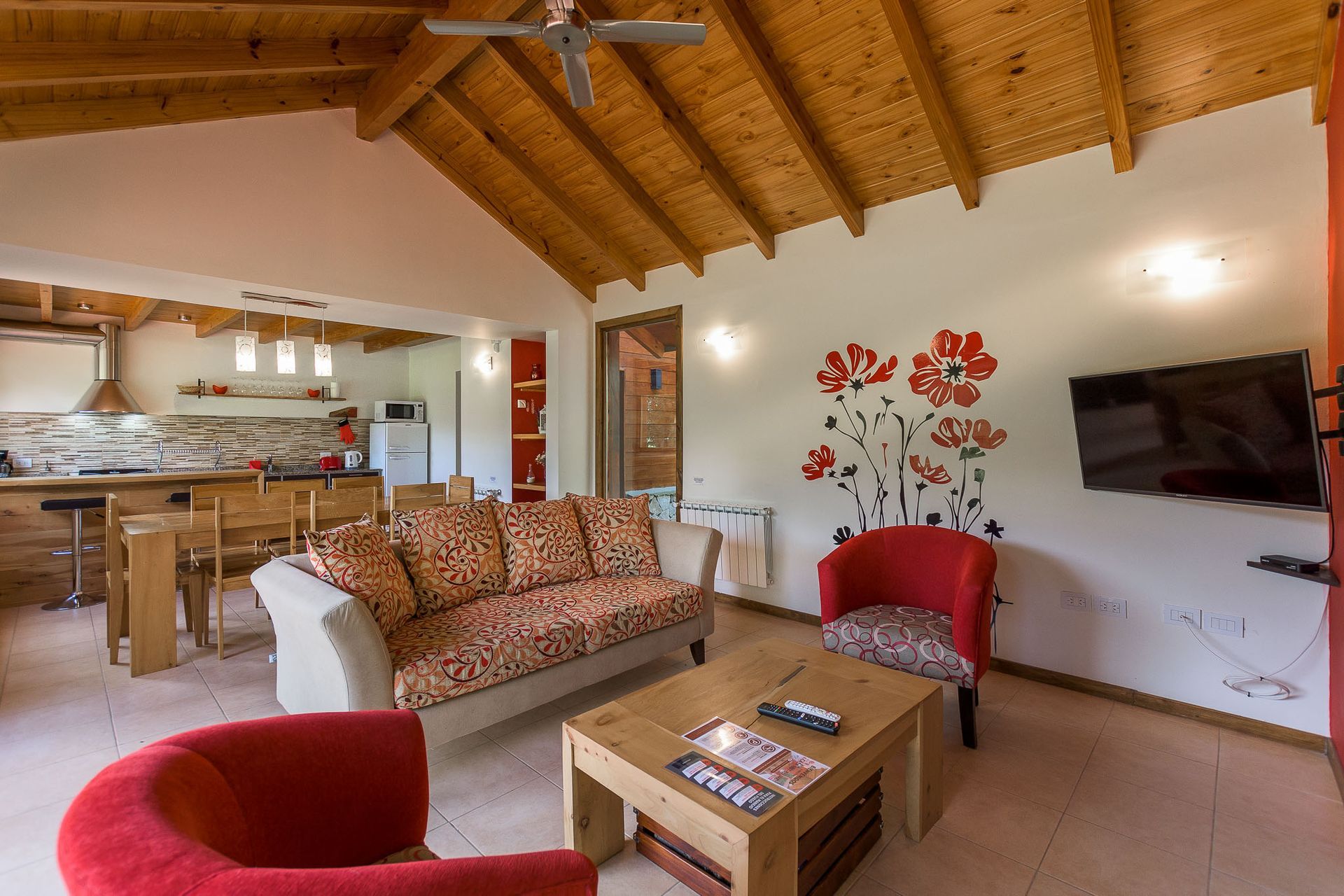 Una sala de estar con un sofá estampado, dos sillones rojos, una mesa de centro de madera y cuadros florales en la pared bajo un techo abovedado.