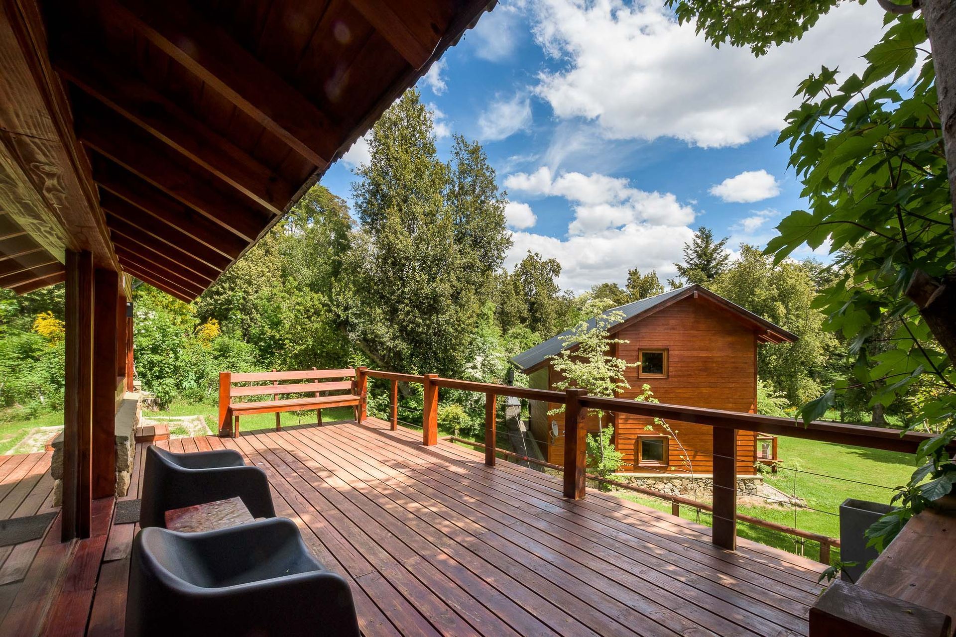 Una terraza de madera con dos sillas oscuras ofrece vistas a una cabaña rústica de troncos rodeada de árboles bajo un cielo azul brillante.