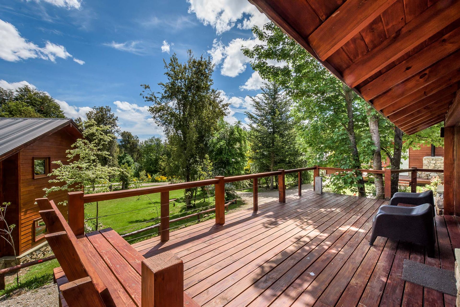 Una terraza de madera con asientos integrados y dos sillas con vistas a un jardín exuberante y soleado bajo un cielo azul parcialmente nublado.