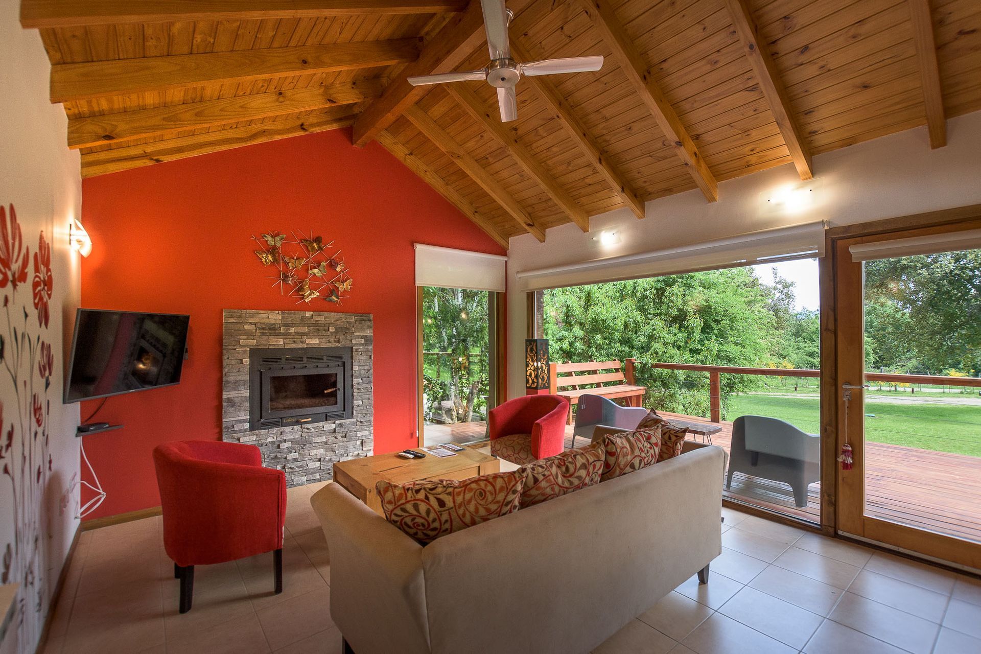 Un salón luminoso con una pared decorativa de color rojo, chimenea de piedra, techo abovedado de madera y vistas a un jardín verde.