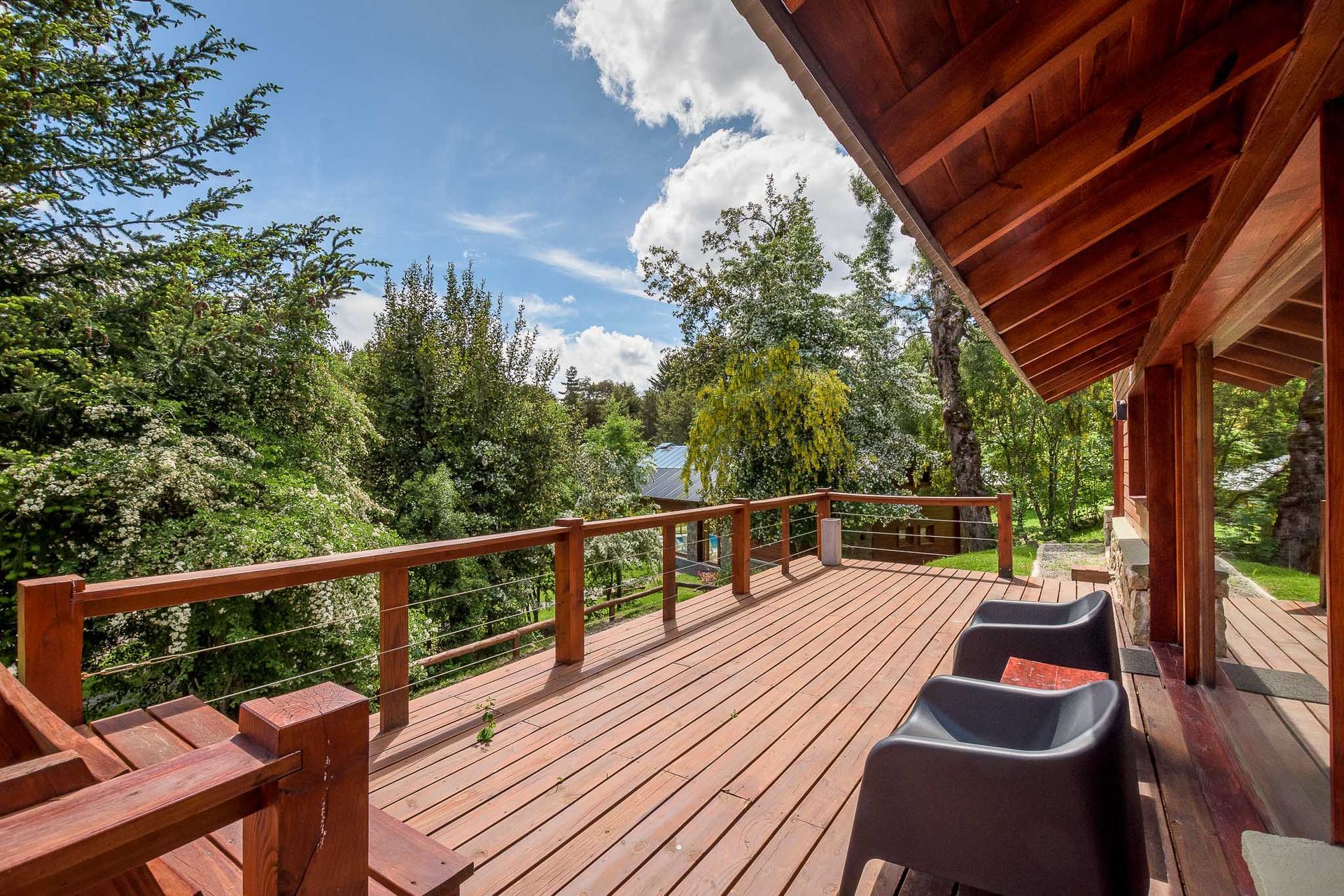 Una terraza de madera con dos sillas oscuras con vistas a un frondoso bosque verde bajo un cielo parcialmente nublado.