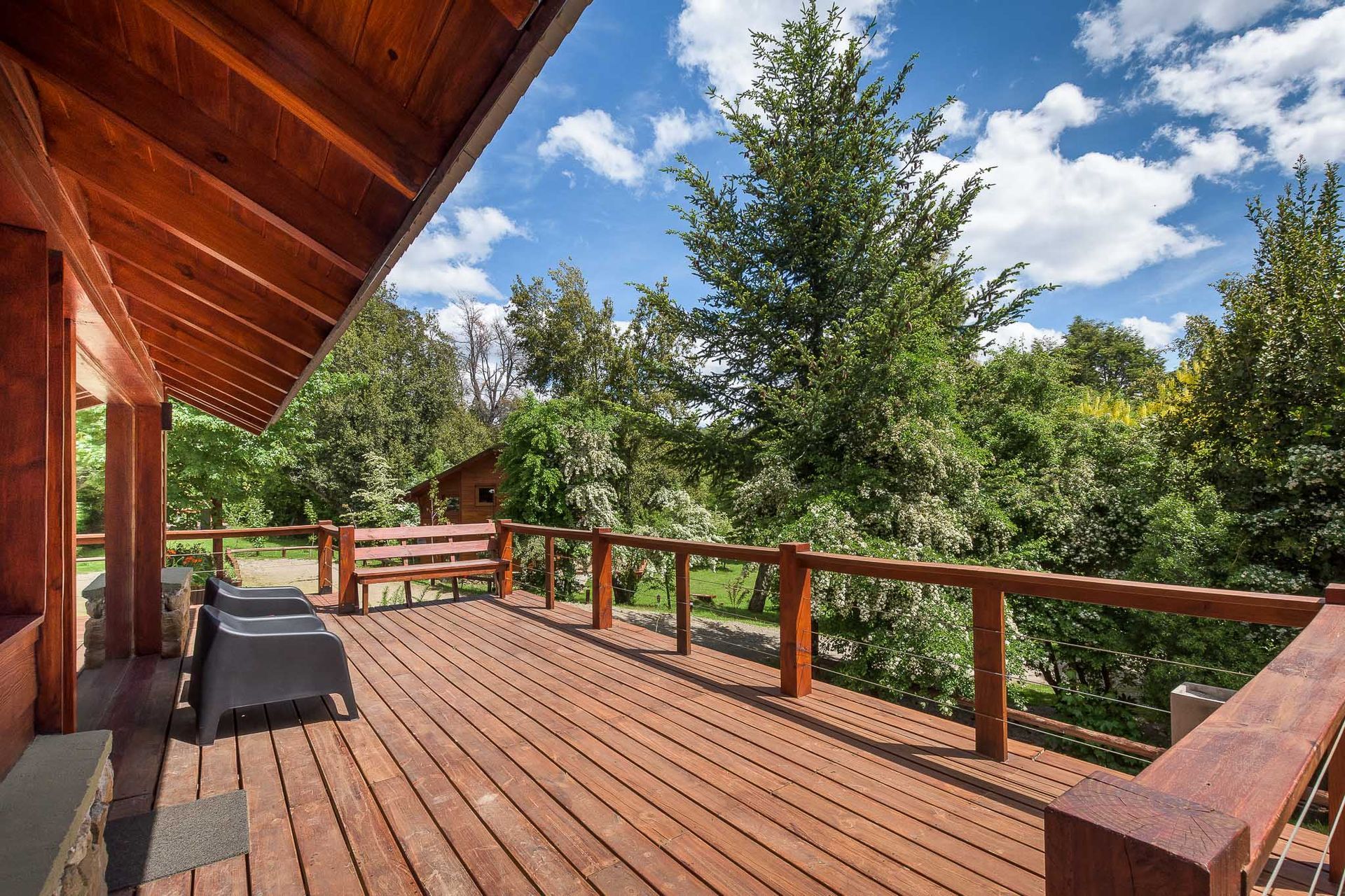 Una terraza de madera ofrece vistas a un jardín frondoso y verde con árboles altos bajo un cielo azul brillante, parcialmente nublado.