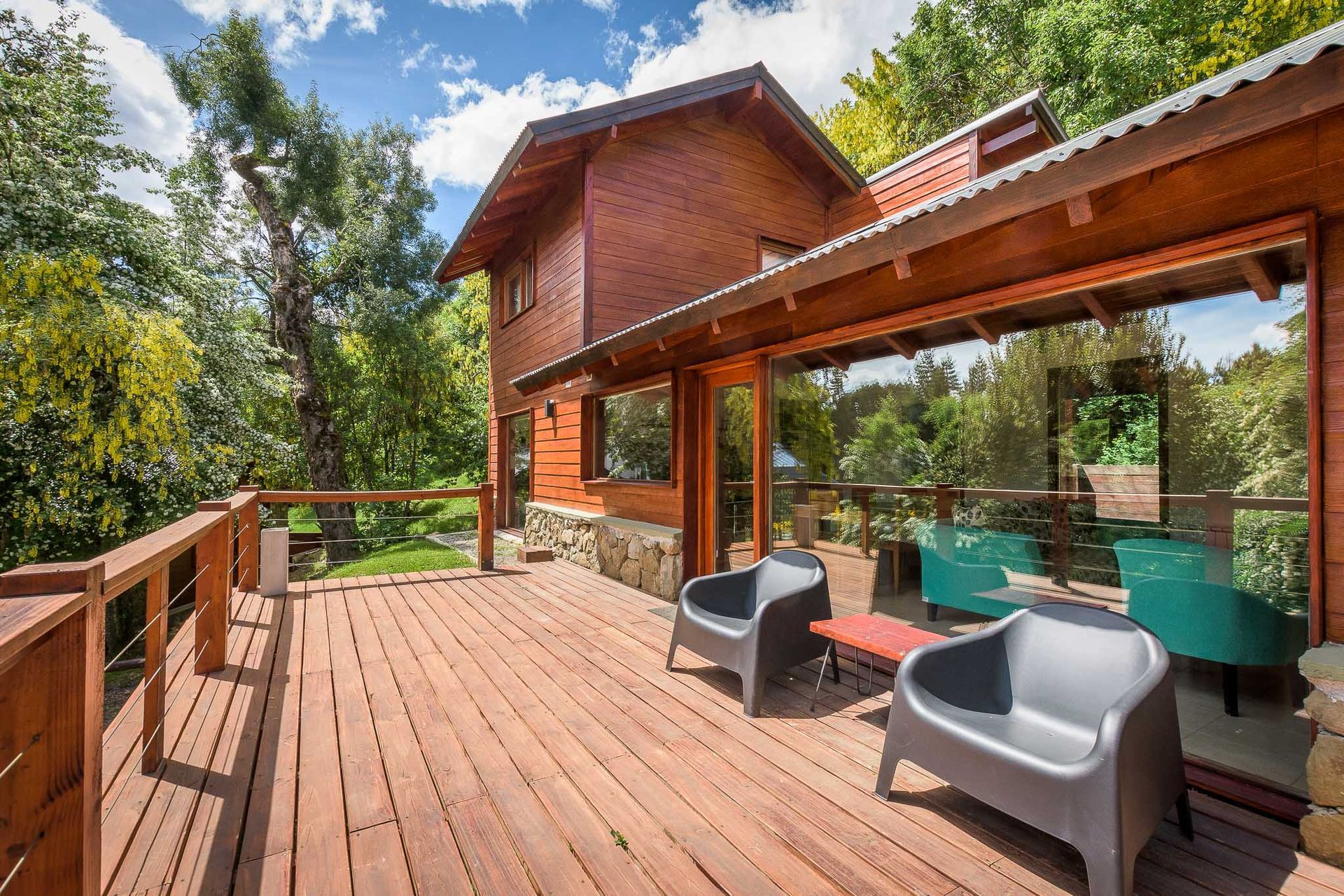 Terraza de madera en el exterior de una cabaña rústica con sillones grises, bajo un techo de metal y rodeada de árboles bajo un cielo azul.