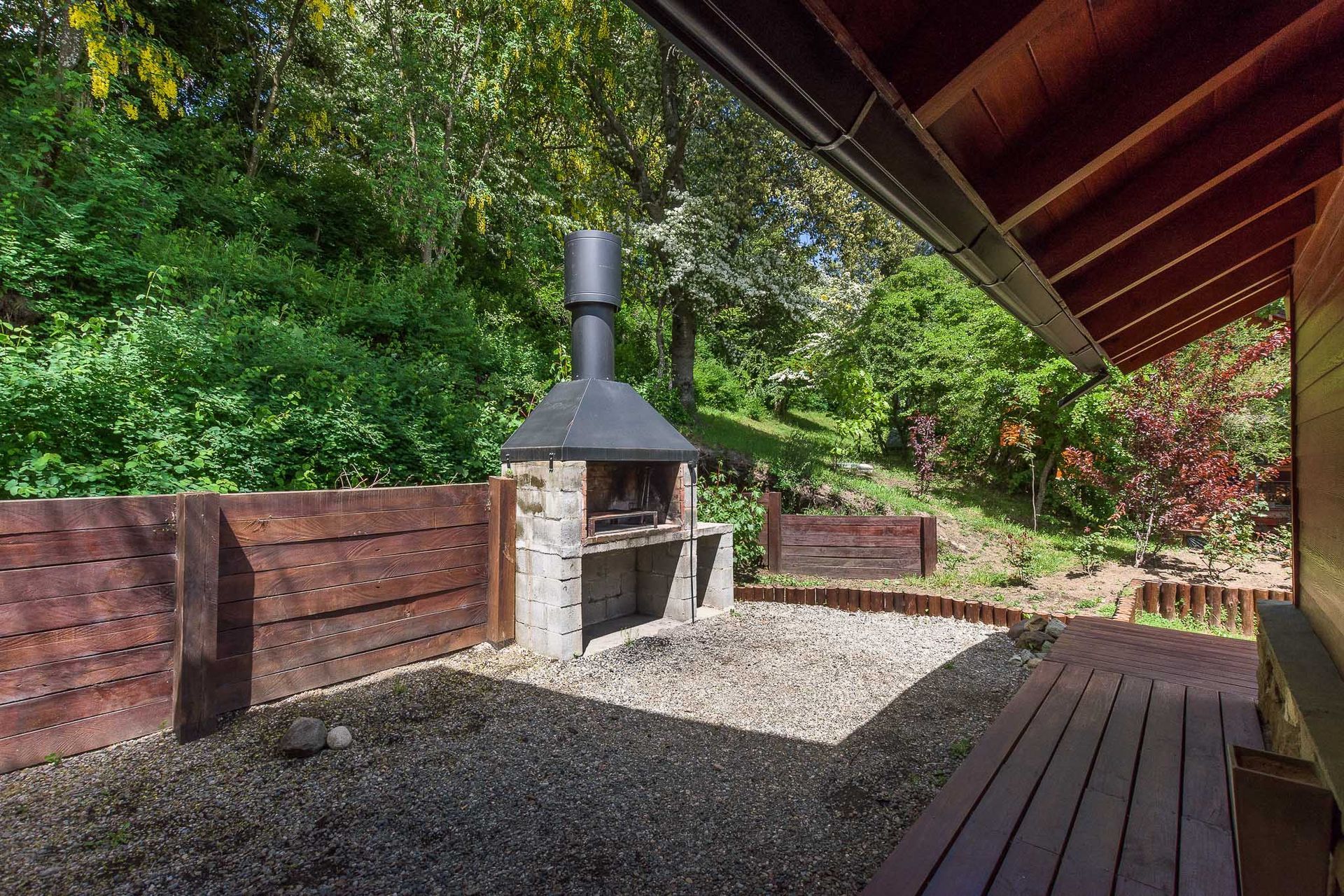 Una chimenea exterior de piedra con una chimenea negra se alza sobre un patio de grava junto a una terraza de madera y un jardín escalonado.