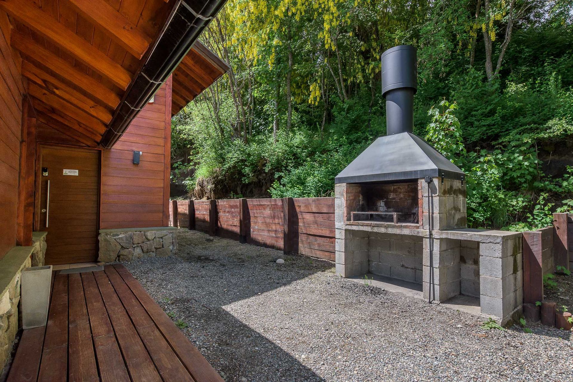 Una barbacoa de piedra al aire libre con chimenea metálica, situada en un patio de grava junto a una cabaña de madera y una valla.