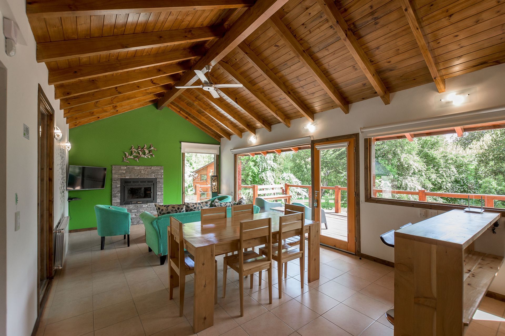 Un luminoso salón-comedor con techos abovedados de madera, una pared decorativa de un verde vibrante y ventanales que van desde el suelo hasta el techo.