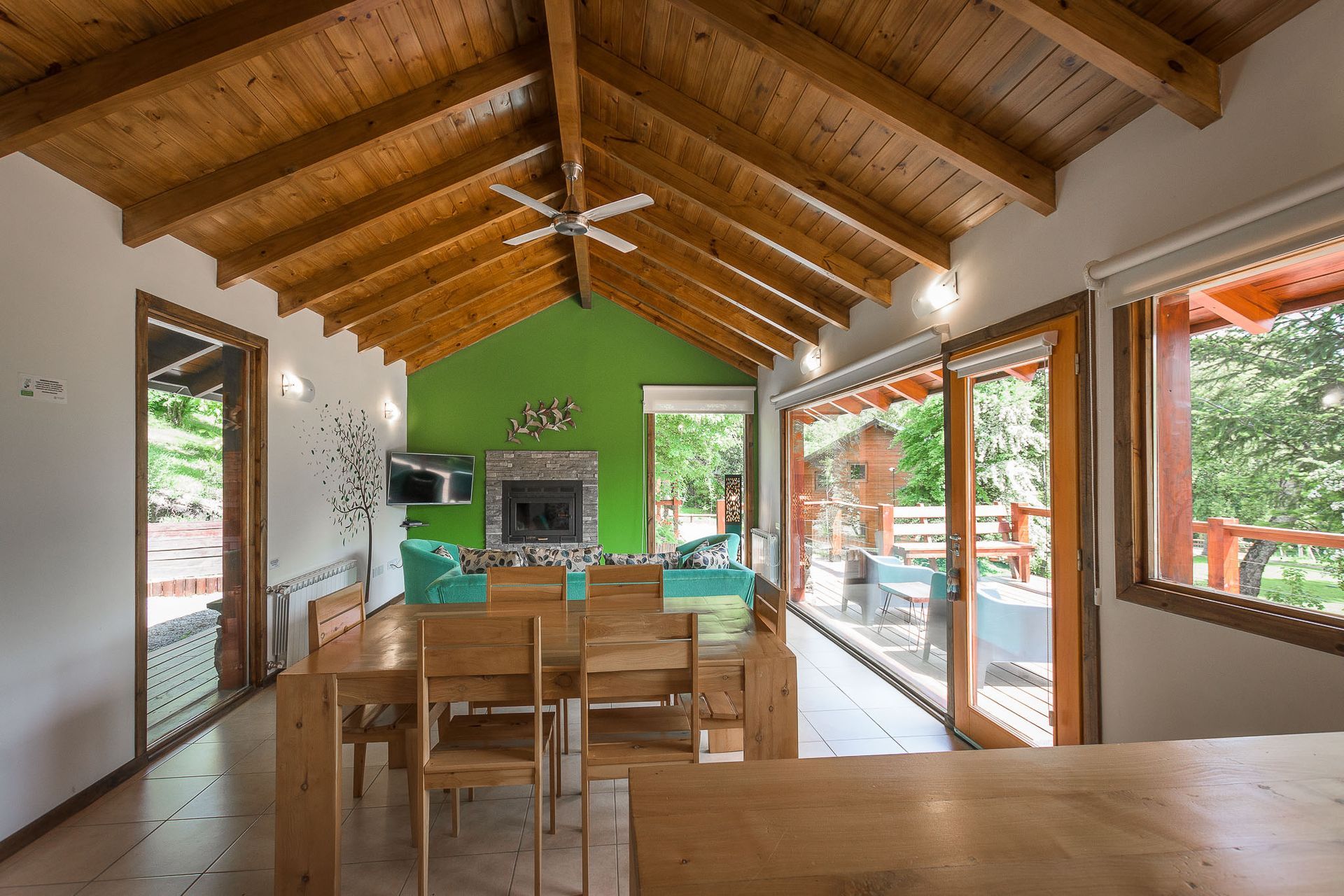 Una sala de estar y comedor con techos altos abovedados de madera, una pared decorativa de color verde, chimenea y puertas de cristal que dan al patio.