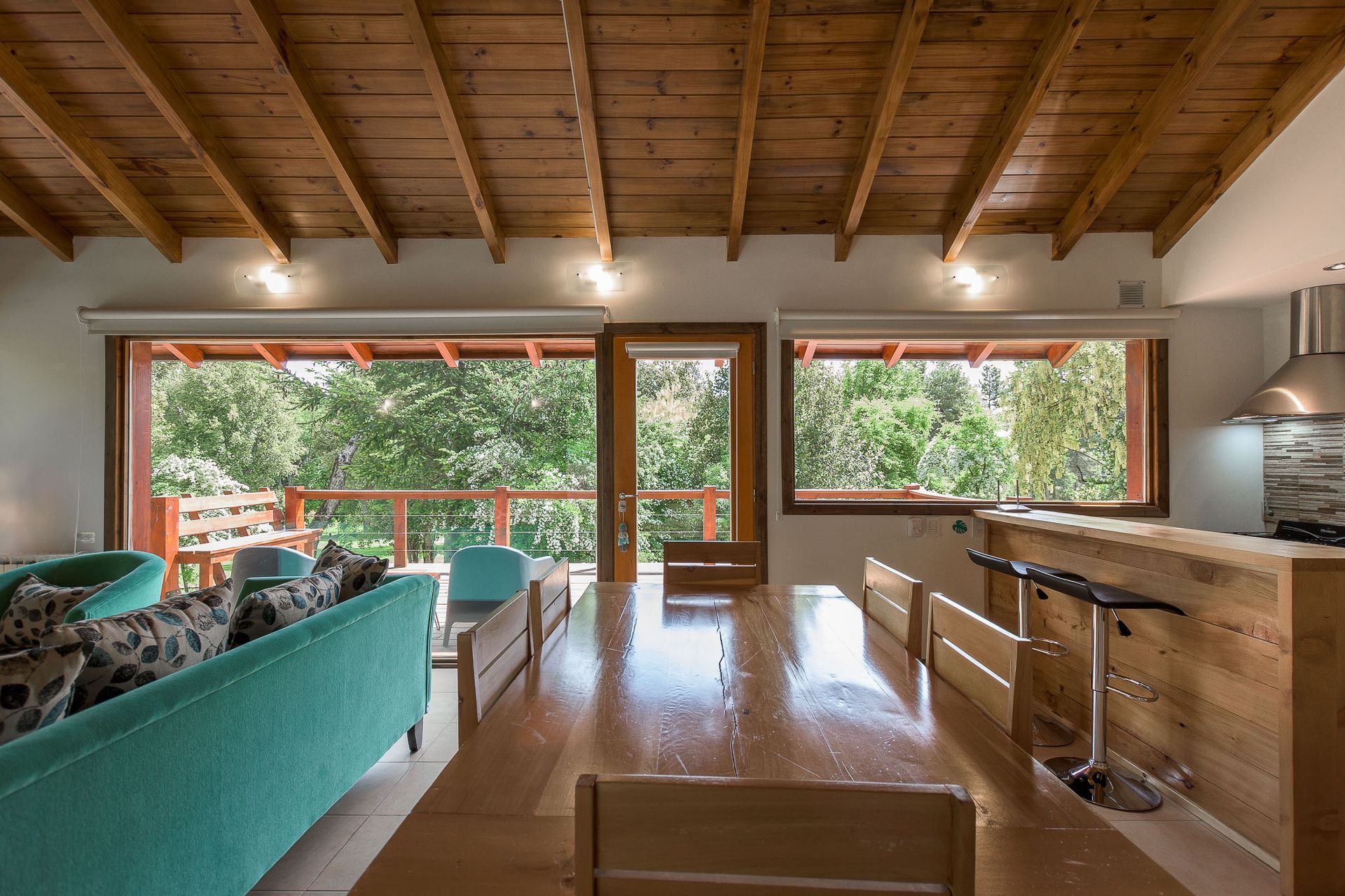 Un espacio diáfano que integra salón y comedor, con un sofá color turquesa, una mesa de comedor de madera y grandes ventanales con vistas al jardín.