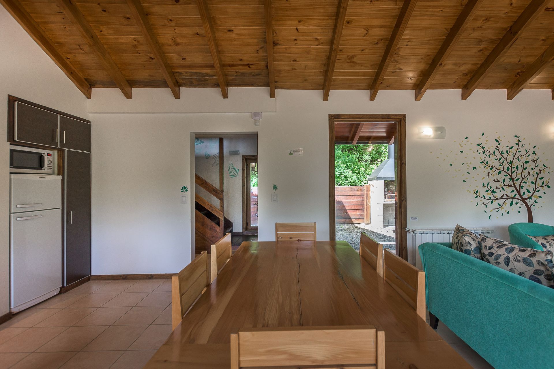 Un comedor rústico con mesa de madera, sillas, nevera, techo abovedado de madera y vistas a una puerta abierta.