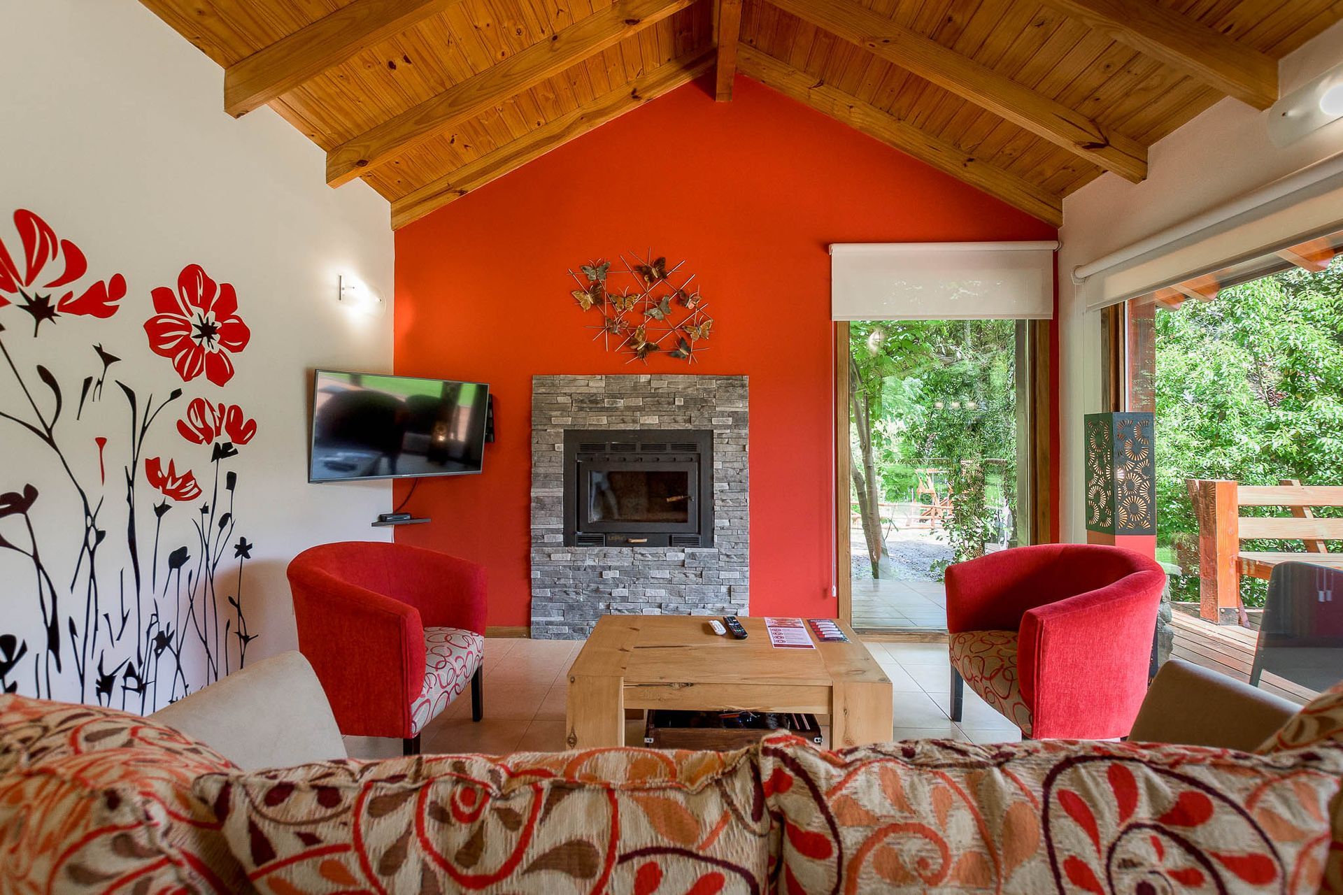 Un salón moderno con una pared decorativa roja, chimenea de piedra, dos sillones rojos, techo de madera y cuadros florales en la pared.