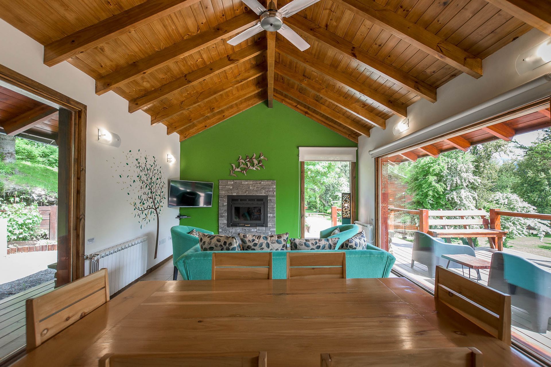 Un luminoso salón con techo abovedado de madera, una pared decorativa de color verde lima, chimenea y sofás de color turquesa.