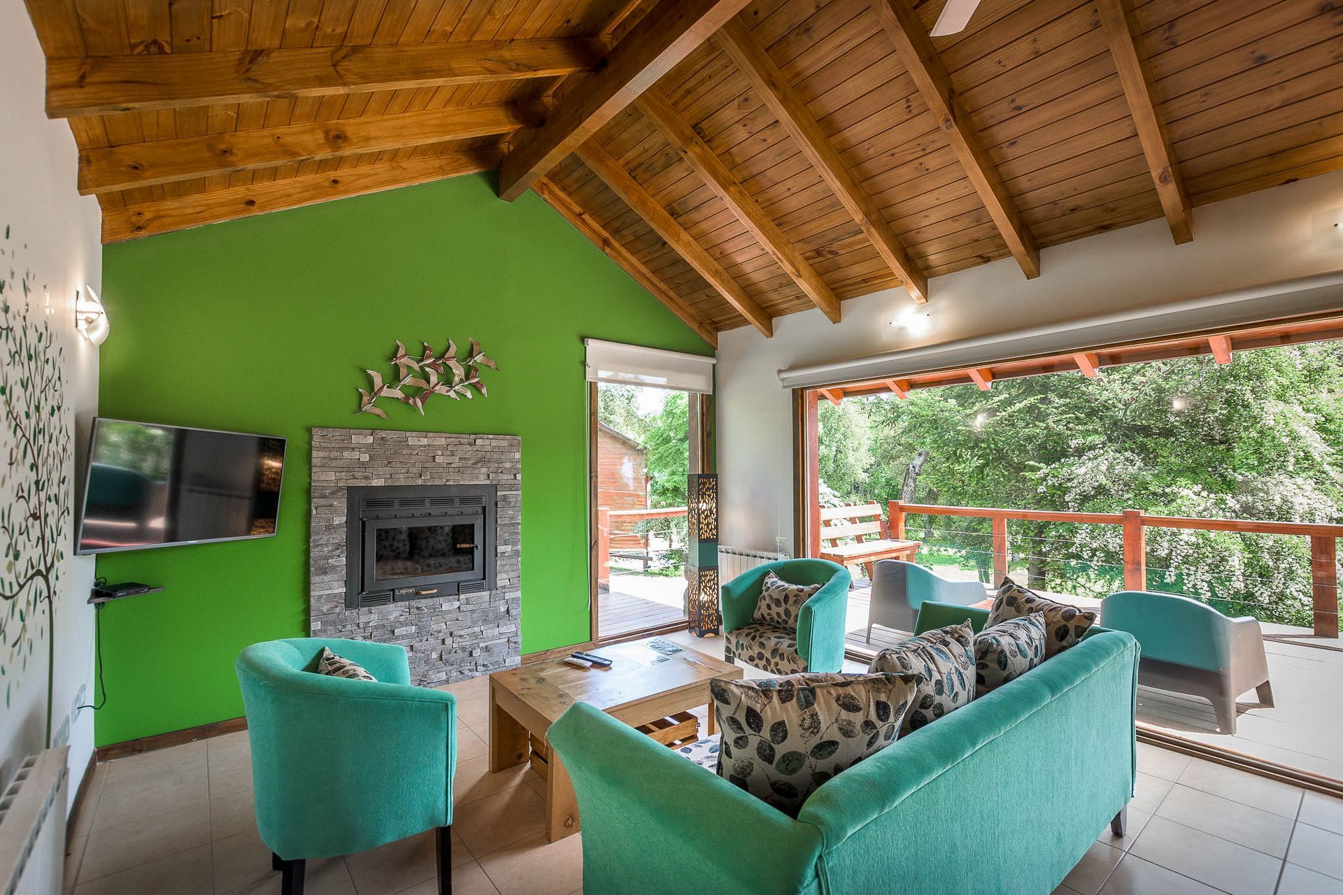 Un luminoso salón con una pared verde, chimenea de piedra, techo abovedado de madera y asientos de color turquesa con vistas a una terraza exterior.
