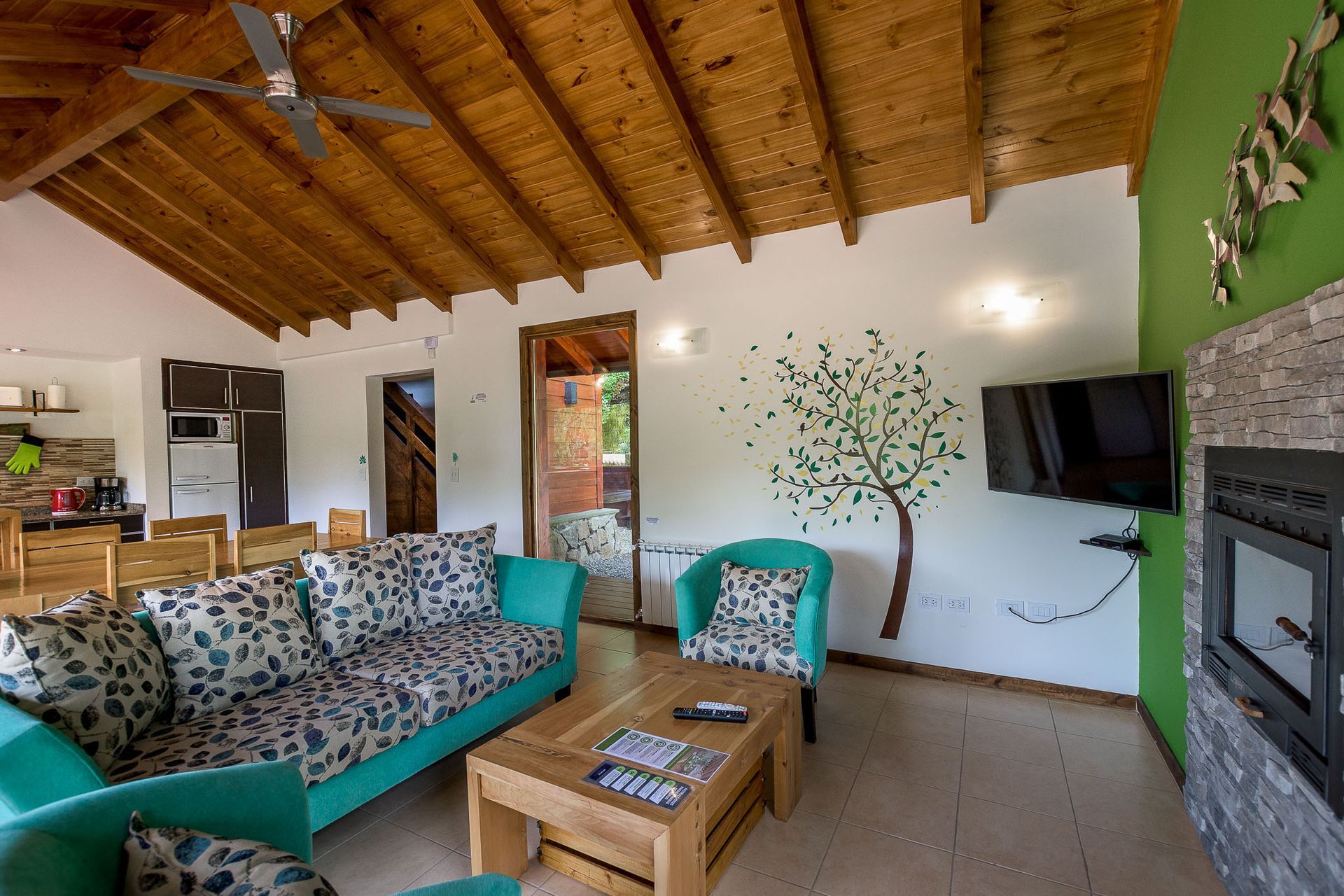 Una sala de estar con un sofá turquesa, una mesa de centro de madera, un sillón, un televisor montado en la pared y un mural de árboles bajo un techo de madera.
