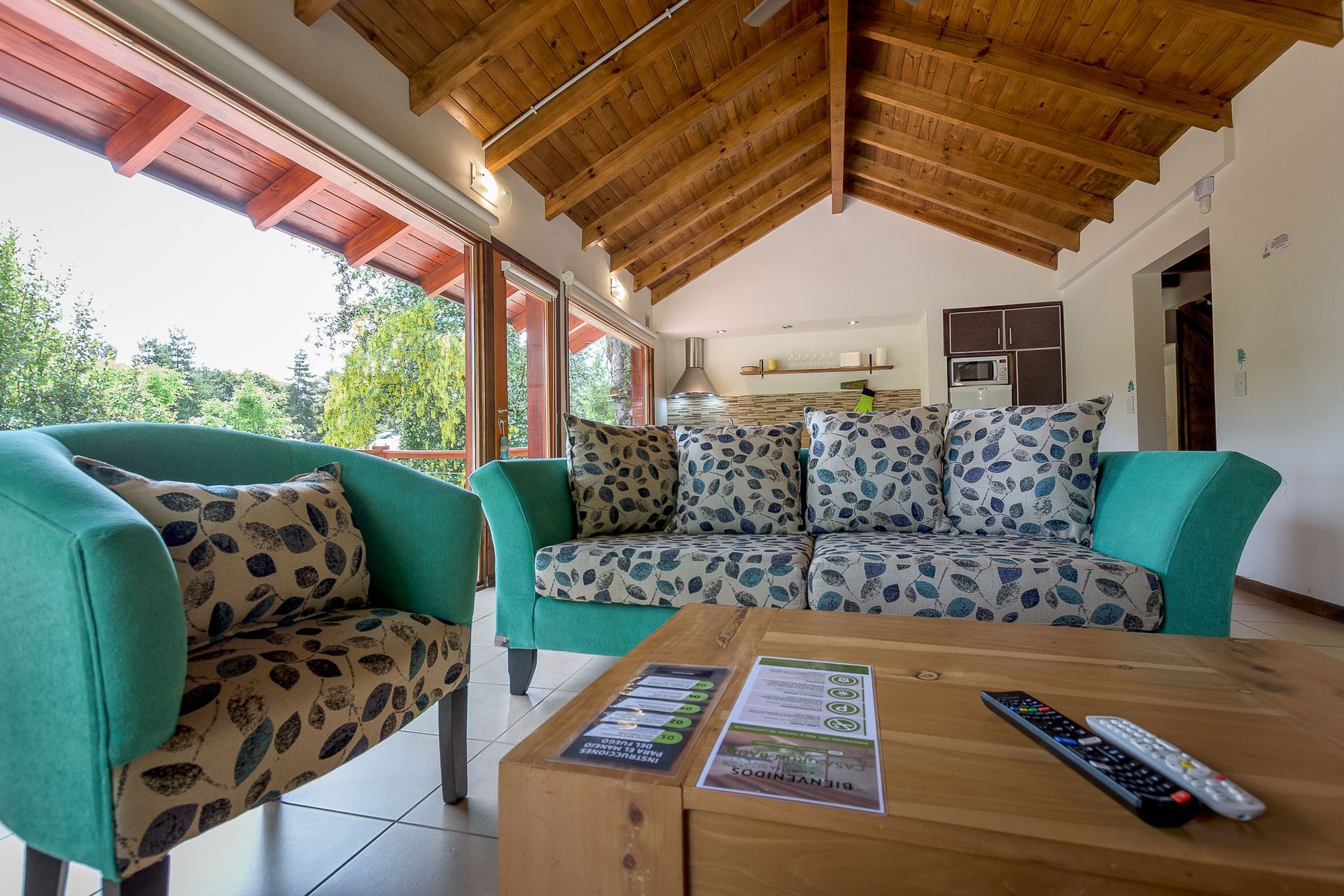 Una acogedora sala de estar con un sofá y un sillón de color turquesa, cojines florales, techo abovedado de madera y una mesa de centro de madera.