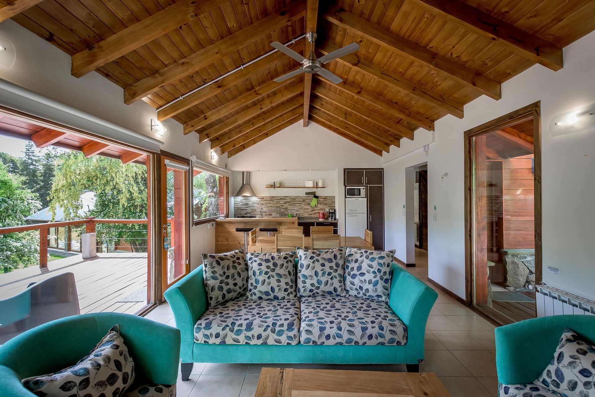 Un luminoso salón con sofá y sillones de color verde azulado, techo abovedado de madera y una cocina abierta que da a una terraza.