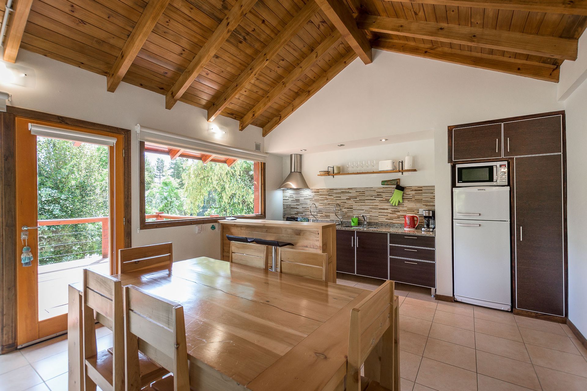 Una cocina y comedor rústicos con mesa de madera, techo con vigas vistas, salpicadero de piedra y nevera blanca.