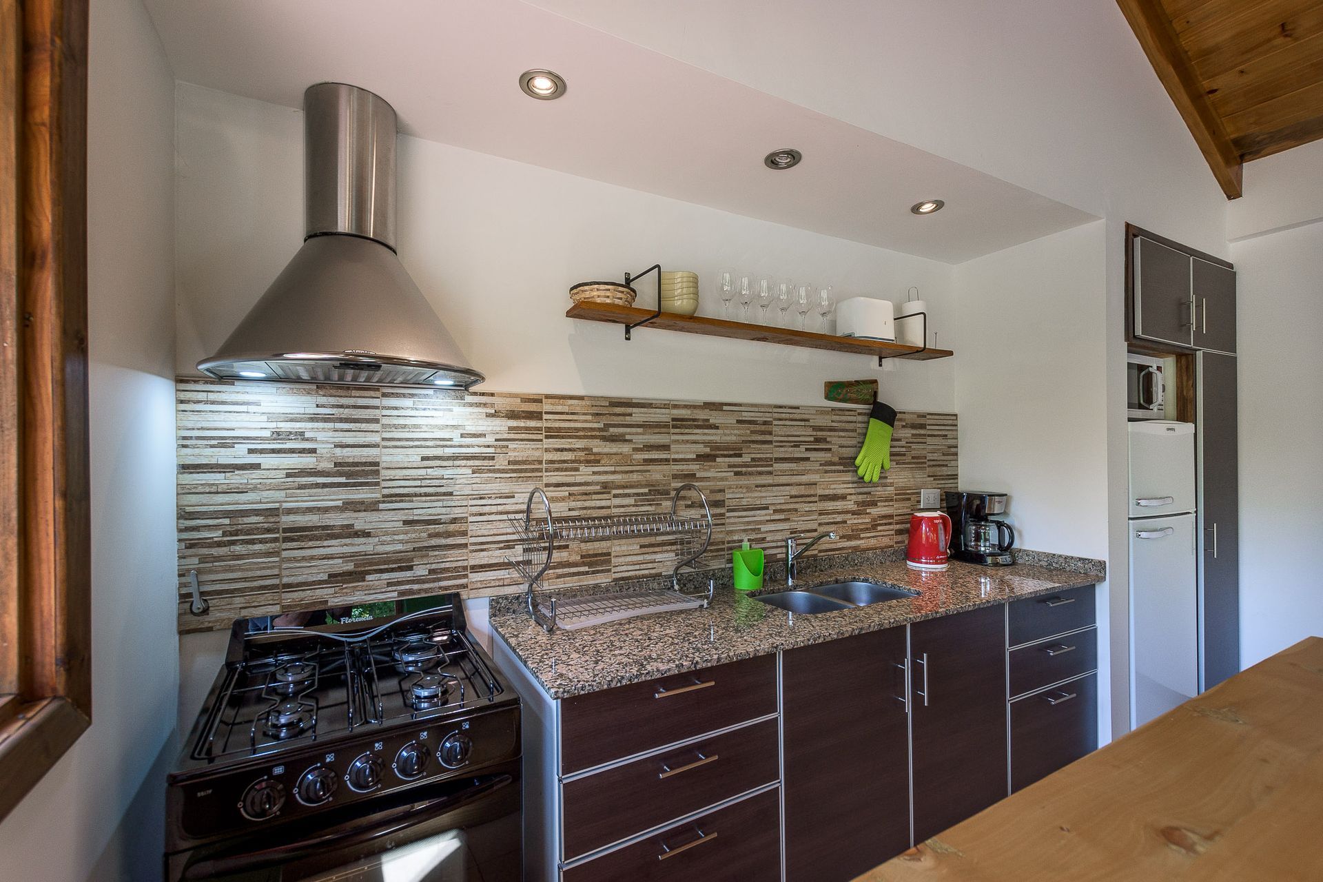 Cocina con encimeras de granito, armarios oscuros, salpicadero de azulejos de piedra, campana extractora de acero inoxidable y frigorífico.