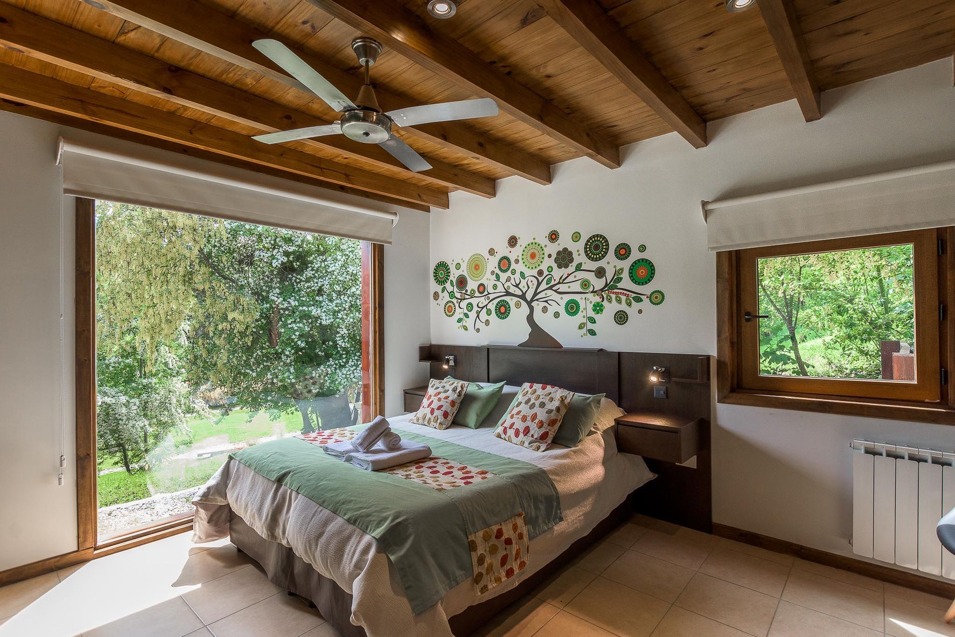 Un dormitorio luminoso con cama de matrimonio, vigas de madera en el techo, ventilador de techo, grandes ventanales y un mural decorativo con un árbol en la pared.