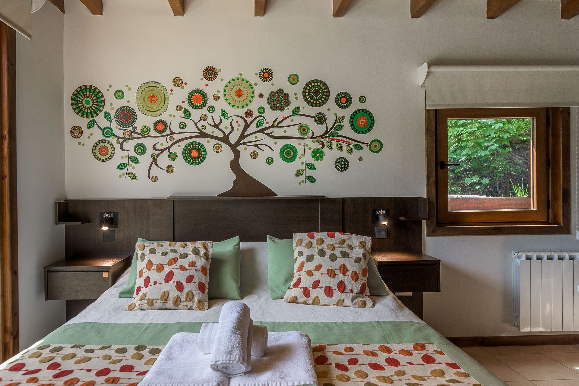 Un dormitorio con una cama doble, cojines florales y un mural en la pared que representa un árbol estilizado con hojas verdes, naranjas y rojas.