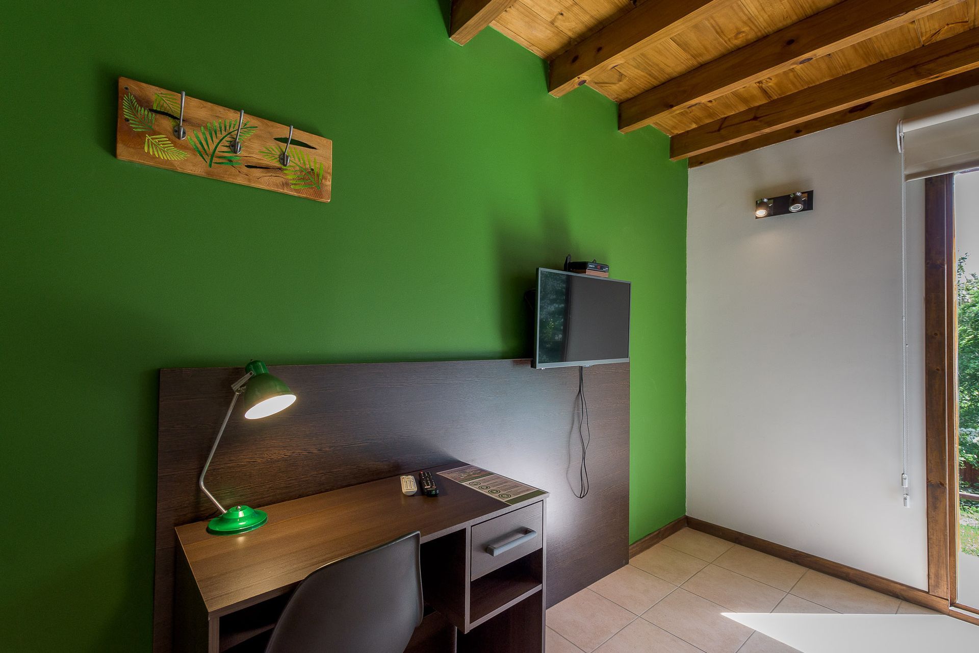 Un escritorio de madera con una lámpara verde y un televisor montado en la pared, frente a una pared de color verde brillante, en una habitación con techo de madera.