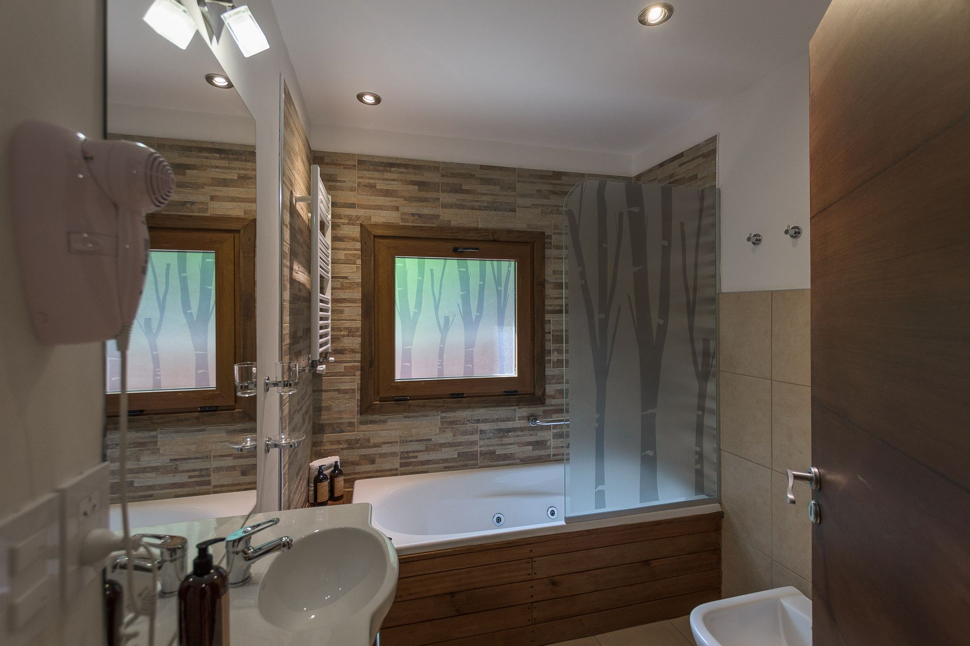 Un baño con lavabo blanco, bañera con mampara de ducha de cristal y una pared con textura de piedra que cuenta con una pequeña ventana.