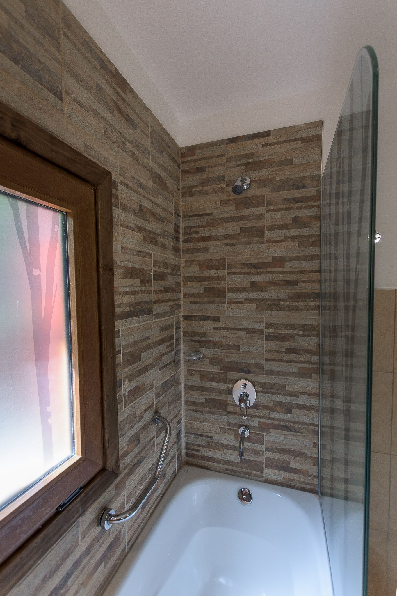 Una bañera con cabezal de ducha, grifería plateada, una barra de apoyo y una mampara de cristal que contrasta con los azulejos de color beige con textura que imitan la piedra.