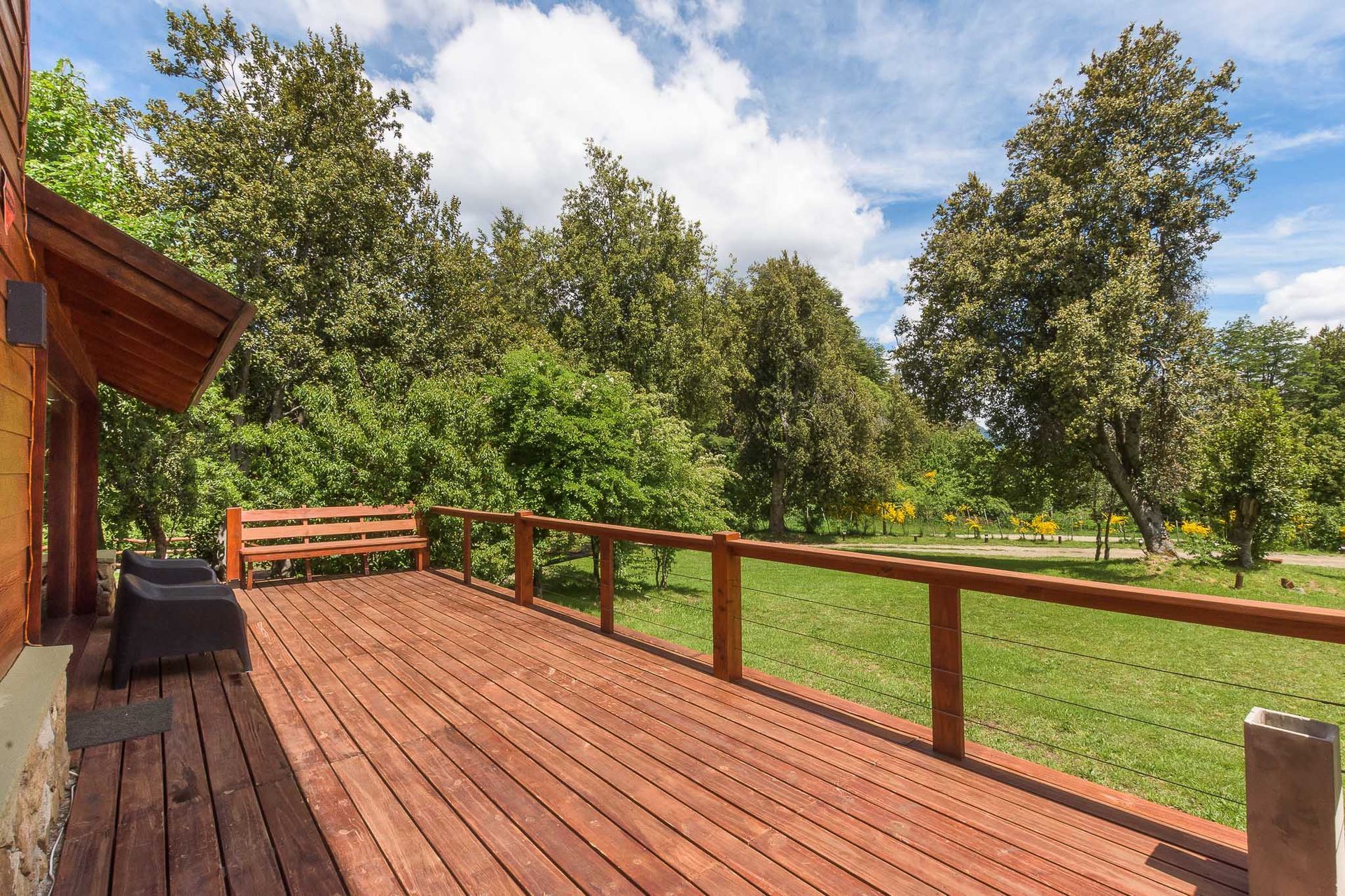 Una terraza de madera con un banco y sillas, con vistas a un jardín cubierto de césped rodeado de frondosos árboles verdes bajo un cielo azul nublado.