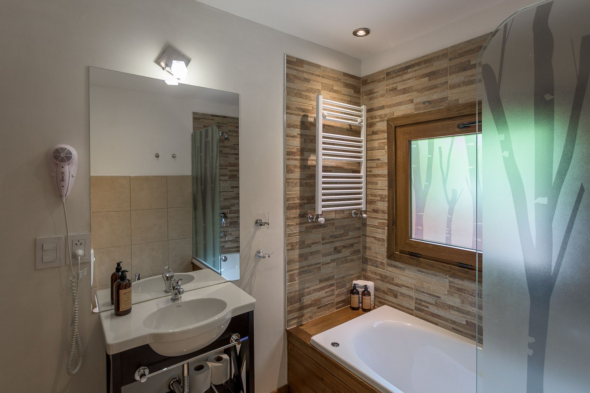 Un baño moderno con tocador blanco con espejo, bañera, toallero eléctrico y azulejos de pared con textura de piedra.