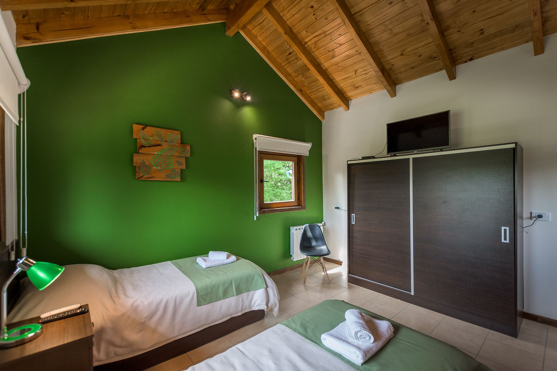 Un dormitorio con dos camas individuales, un gran armario oscuro, una pared decorativa de color verde y un techo de madera inclinado.