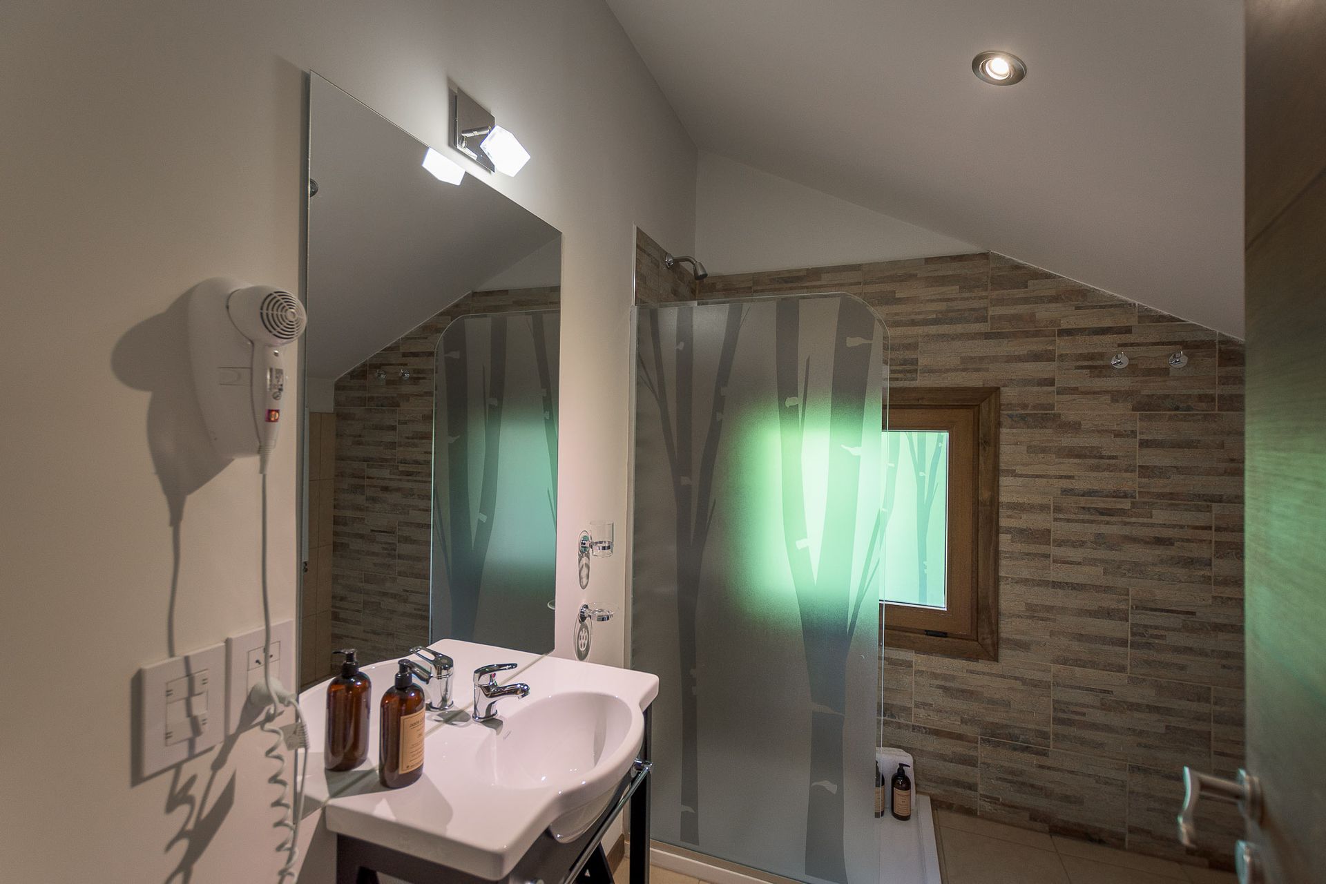 Un baño moderno cuenta con un lavabo blanco, un espejo y una puerta de ducha de vidrio esmerilado con un diseño de árbol sobre una pared alicatada.