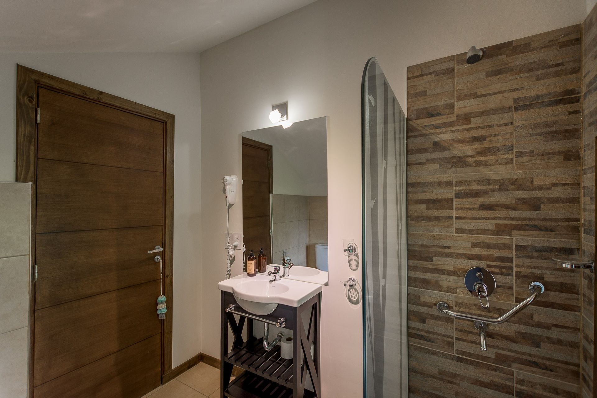 Un moderno baño de hotel con puerta revestida de madera, lavabo blanco con pedestal y espejo iluminado, y ducha alicatada.