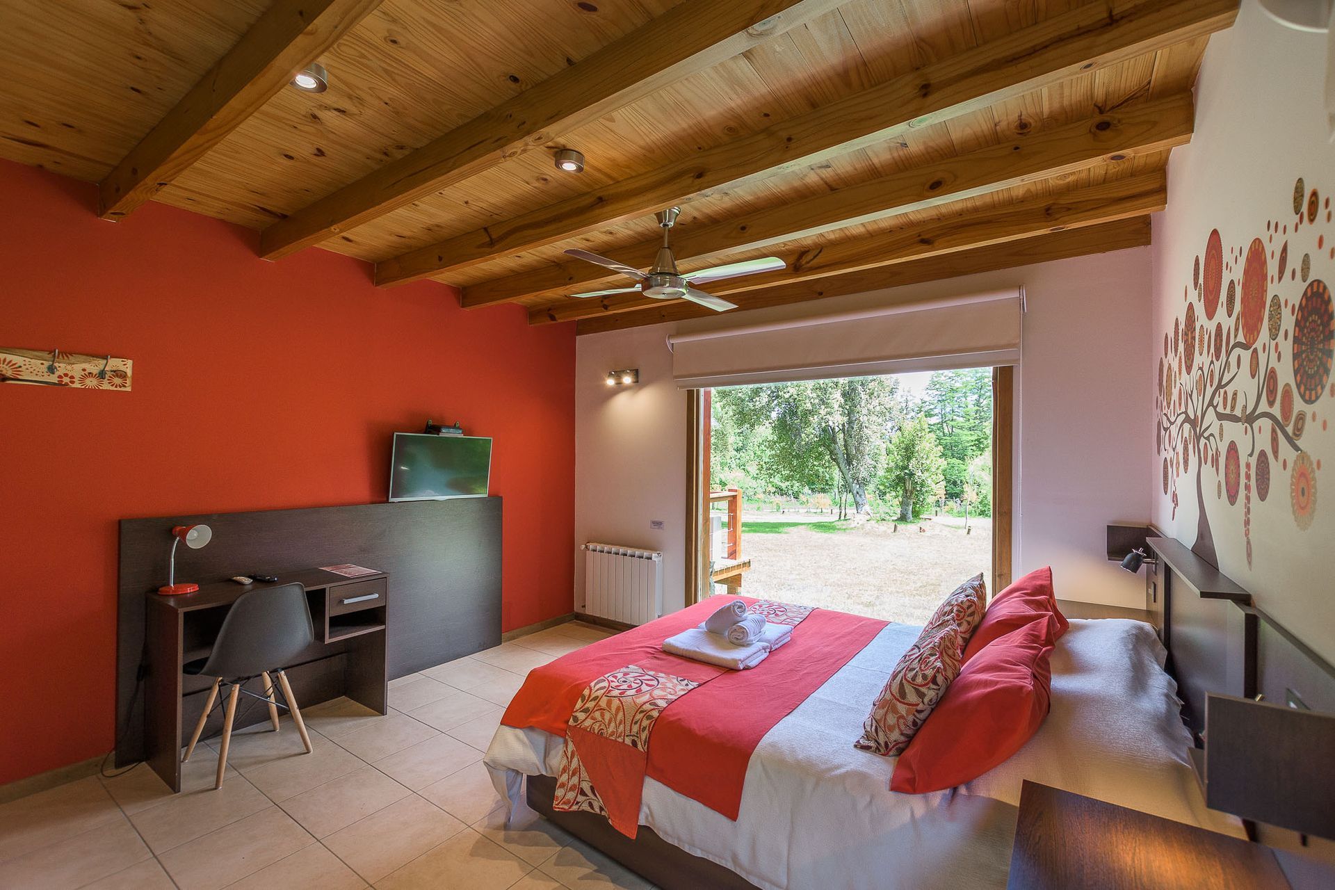 Un dormitorio con una cama con ropa de cama naranja, un escritorio con silla y grandes ventanales con vistas a una zona de césped.