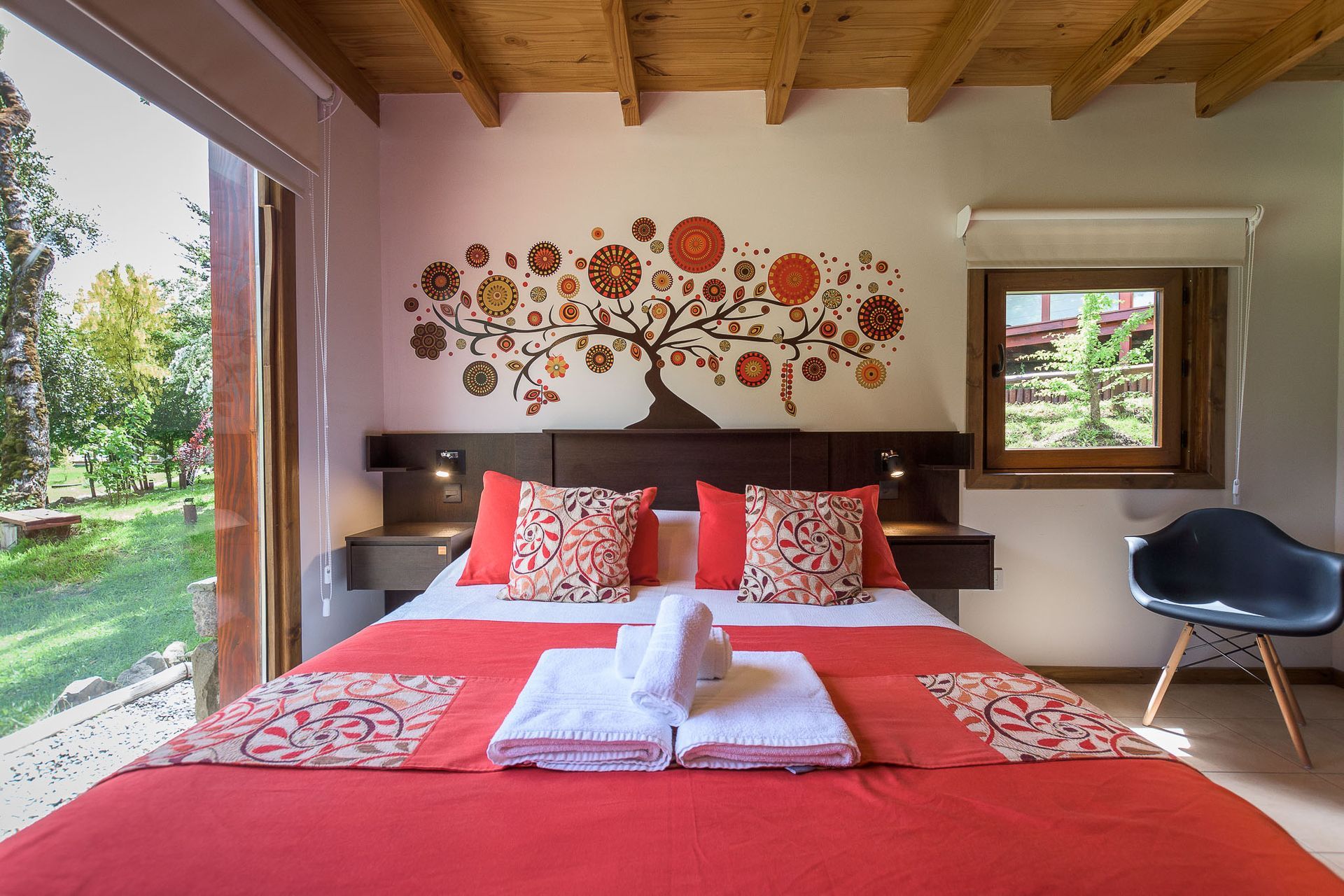 Un dormitorio con una cama roja, cojines estampados, un mural floral en la pared, vigas de madera en el techo y vistas a un jardín.