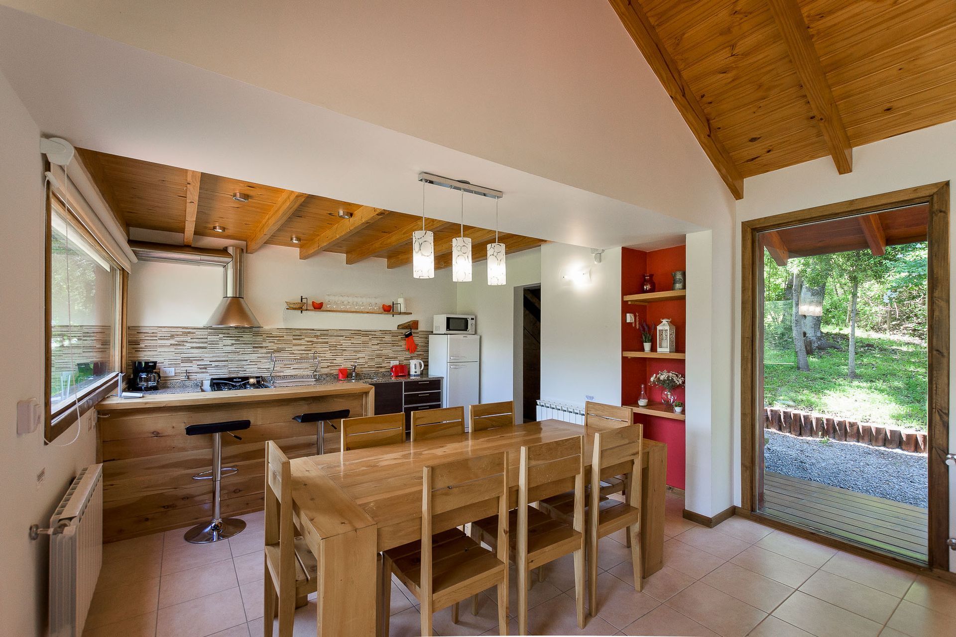 Una cocina y comedor rústicos con mesa de madera, sillas, una isla y una puerta abierta que da a un jardín exterior con césped.