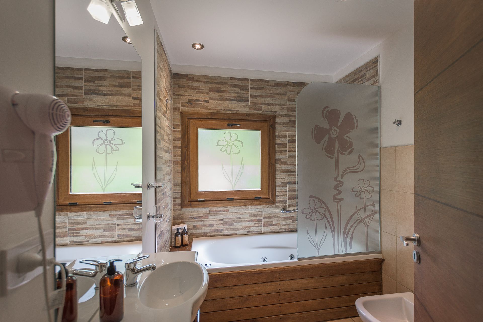 Un baño con bañera de hidromasaje con una mampara de cristal decorativa grabada con flores, detalles en madera y paredes revestidas de azulejos de piedra.