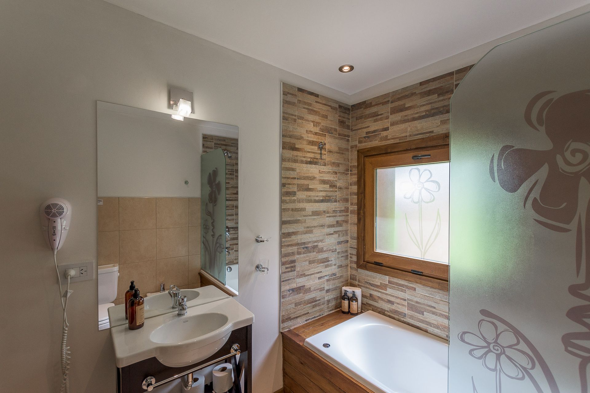 Baño moderno con lavabo, espejo y bañera contra una pared revestida de azulejos de piedra con ventana.