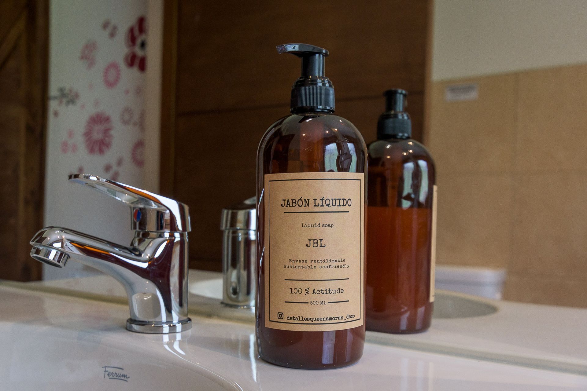 Sobre una encimera blanca de baño, junto a un grifo cromado brillante, reposan botellas de jabón de color ámbar.