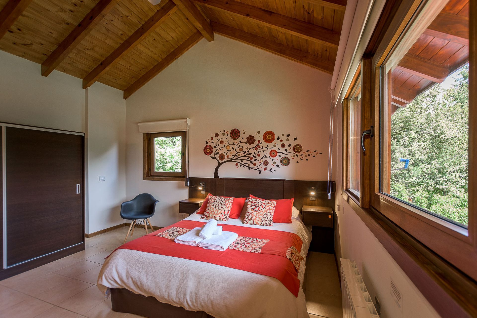 Un dormitorio acogedor con una colcha roja, un vinilo decorativo de un árbol en la pared, un techo alto de madera y ventanas con vistas a árboles verdes.