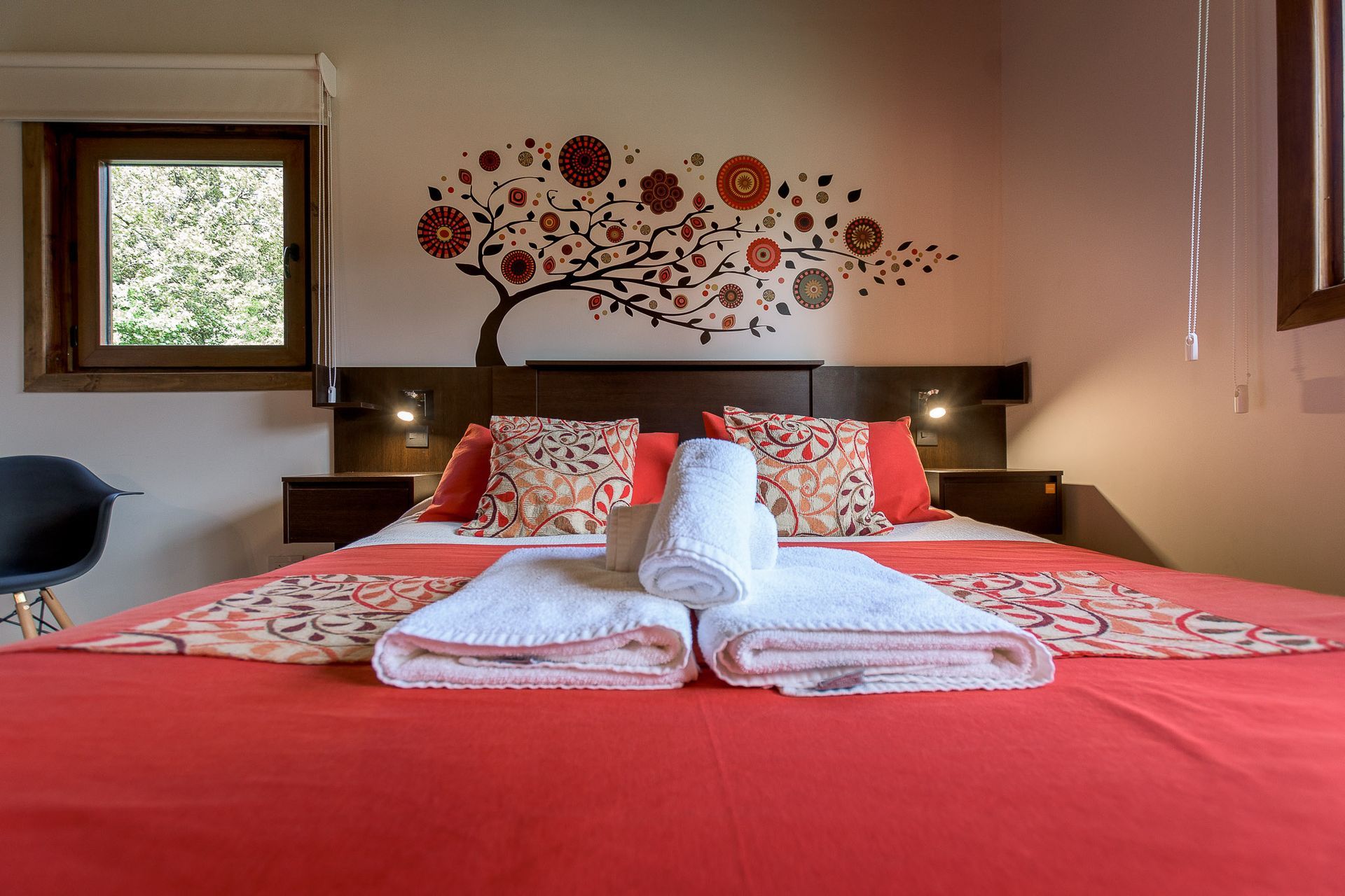 Una cama de hotel con una colcha de color rojo vibrante, cojines decorativos con motivos florales y toallas blancas dobladas frente a un mural con temática de árboles.