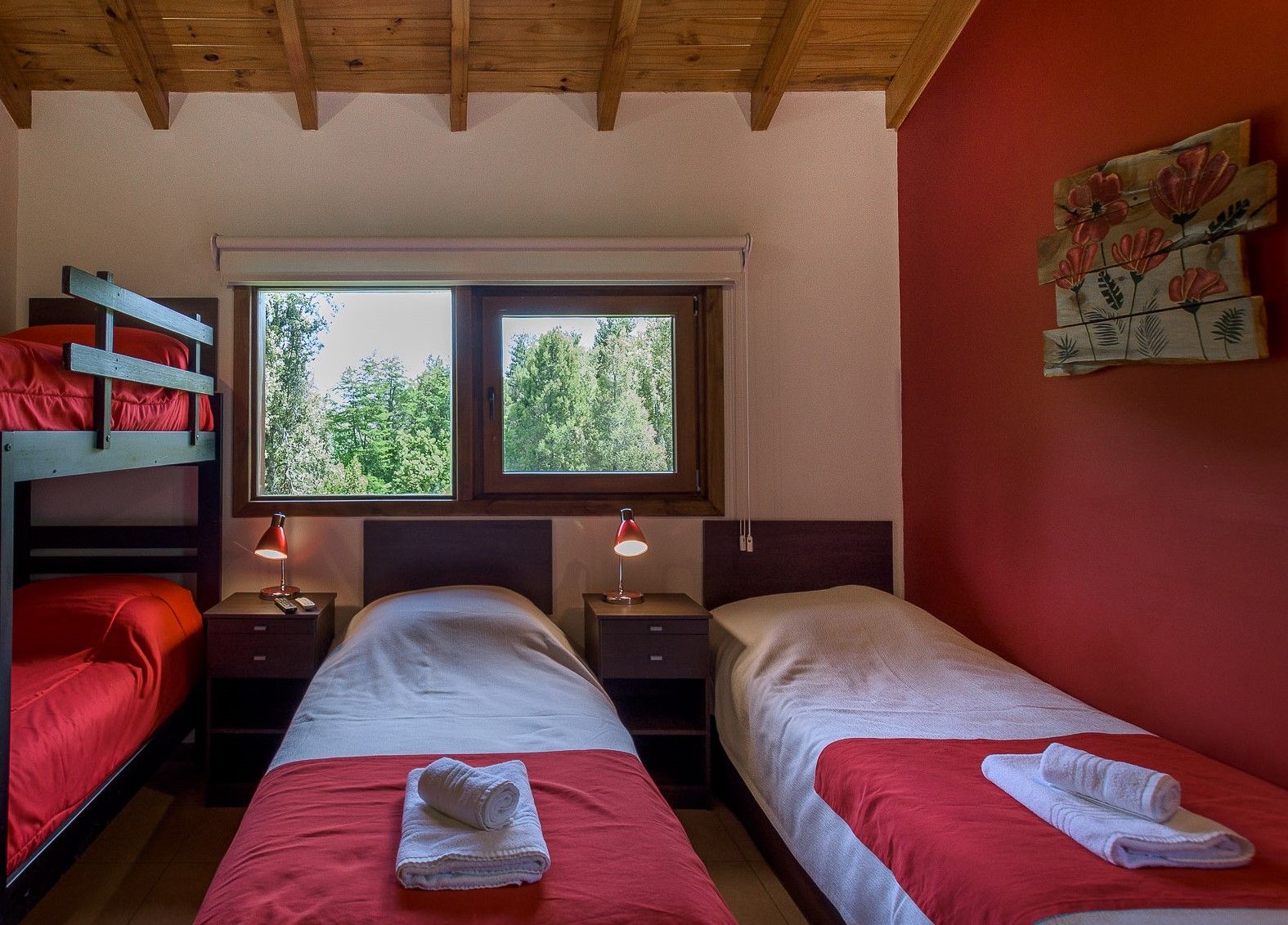 Un dormitorio con una litera, dos camas individuales con sábanas rojas y toallas blancas, mesitas de noche y una pared decorativa roja.