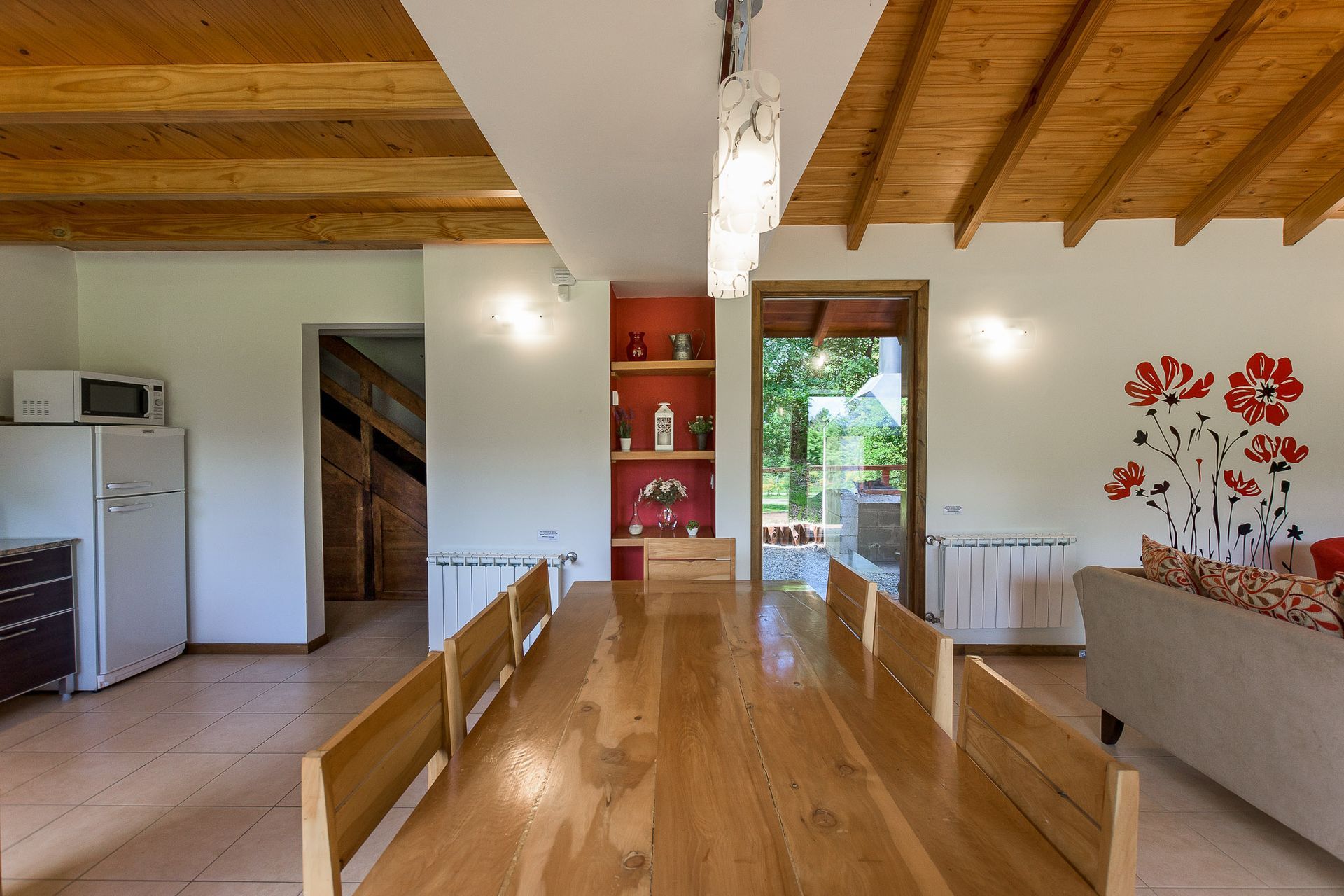 Un comedor rústico con una larga mesa de madera, paredes blancas, vigas de madera a la vista en el techo y un nicho decorativo rojo en la pared.