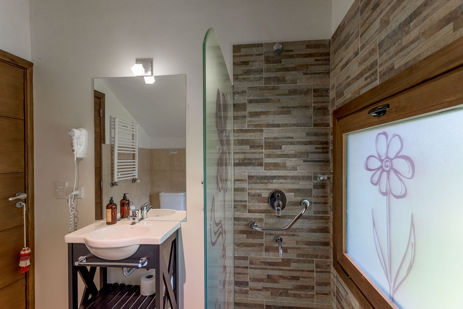 Baño de hotel moderno con lavabo de pedestal, secador de pelo de pared, cabina de ducha de cristal y ventana floral esmerilada.