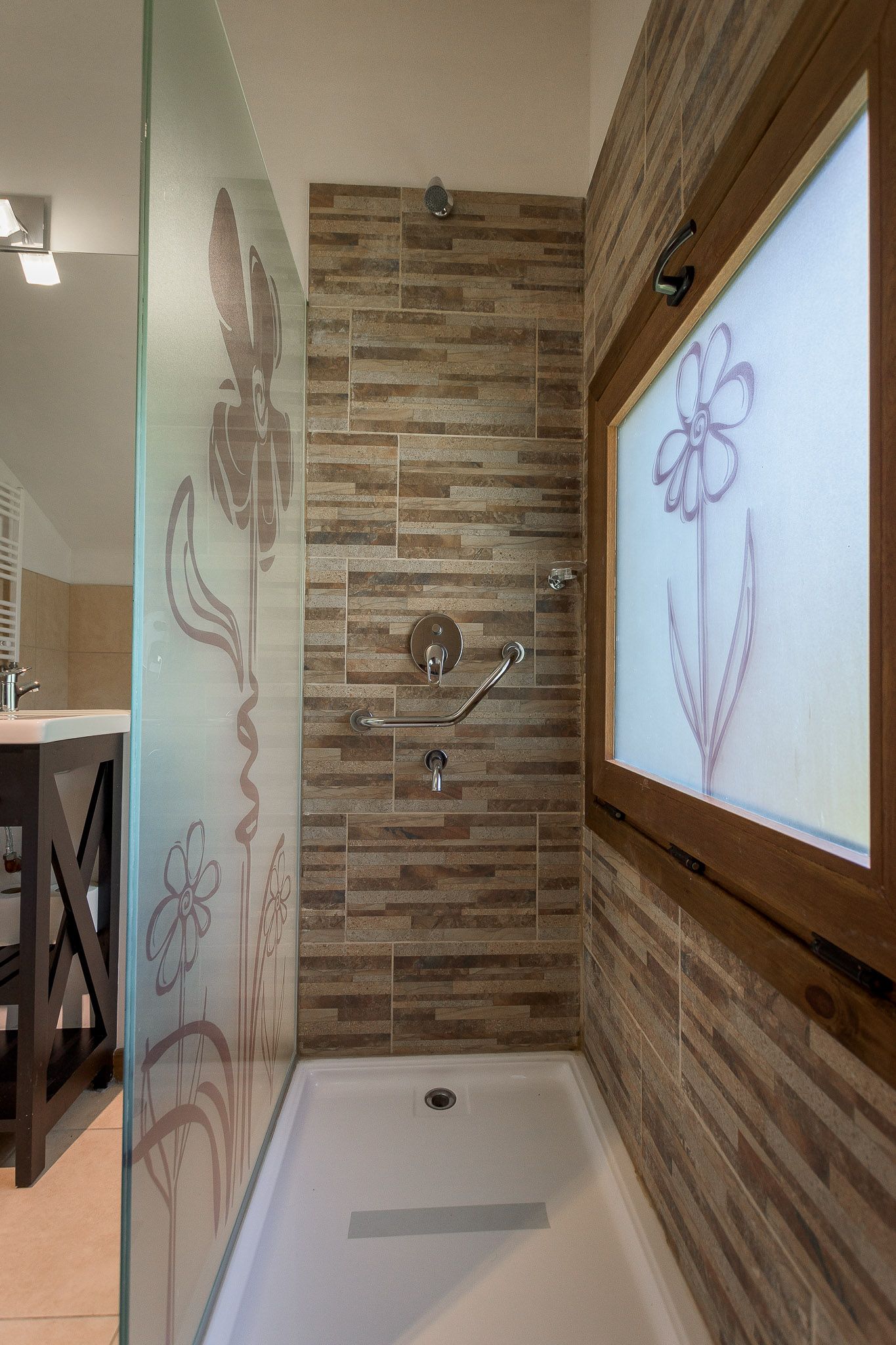 Una ducha a ras de suelo con paredes de piedra apilada, una mampara de cristal con grabados florales y una ventana esmerilada con diseño floral.