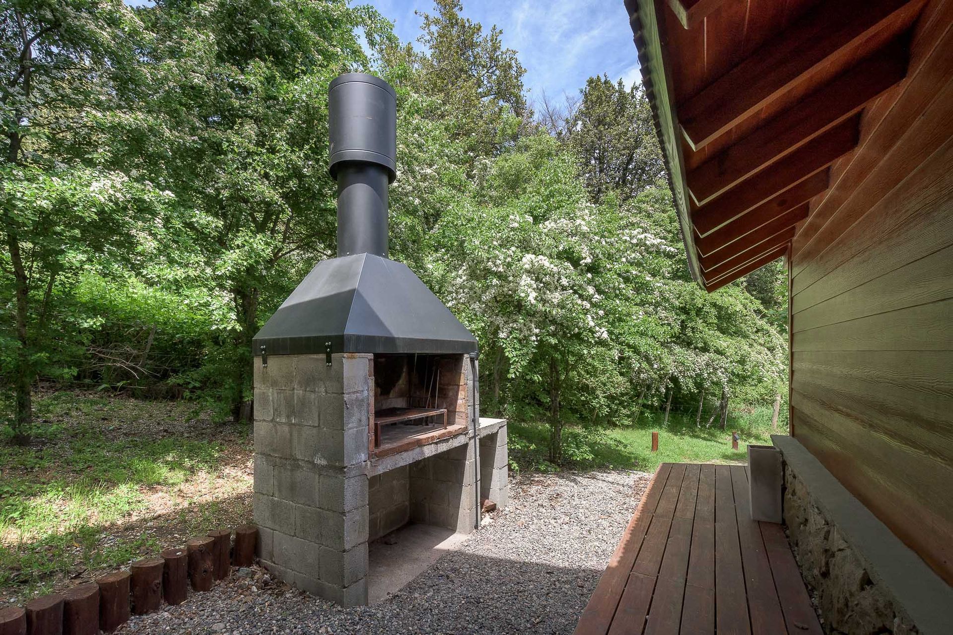Una parrilla de piedra para exteriores con chimenea metálica se alza sobre un patio de grava junto a una terraza de madera y árboles.
