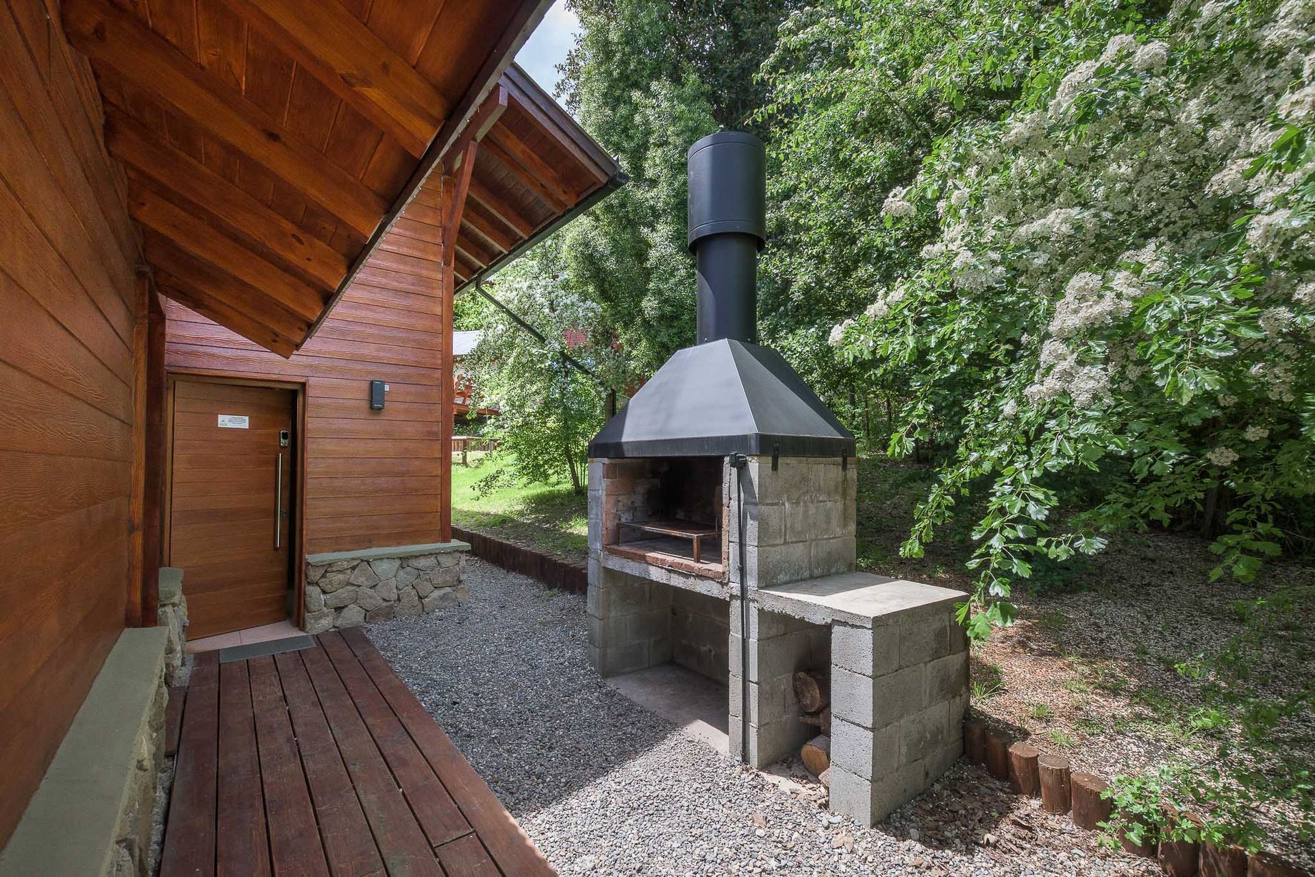 Una parrilla de piedra al aire libre con una chimenea de metal se encuentra en un patio de grava junto a un edificio de madera y frondosos árboles verdes.
