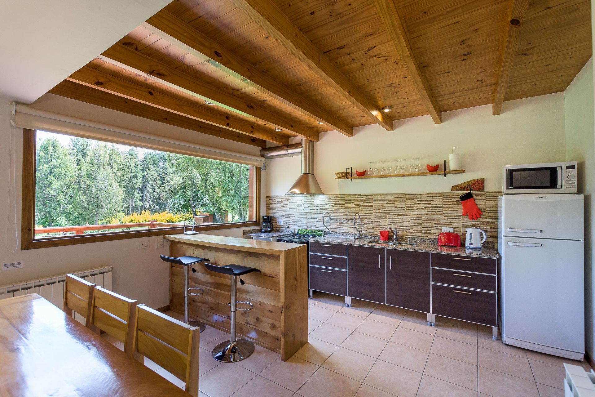 Una cocina rústica con techos de vigas de madera, una barra para desayunar, sillas de madera y un gran ventanal con vistas a los árboles.