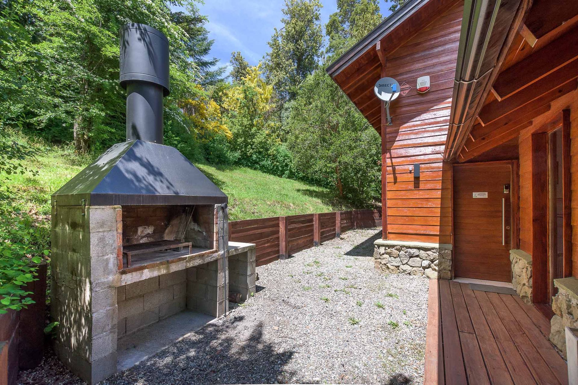 Chimenea de piedra al aire libre junto a una cabaña de madera y un patio de grava rodeado de árboles.