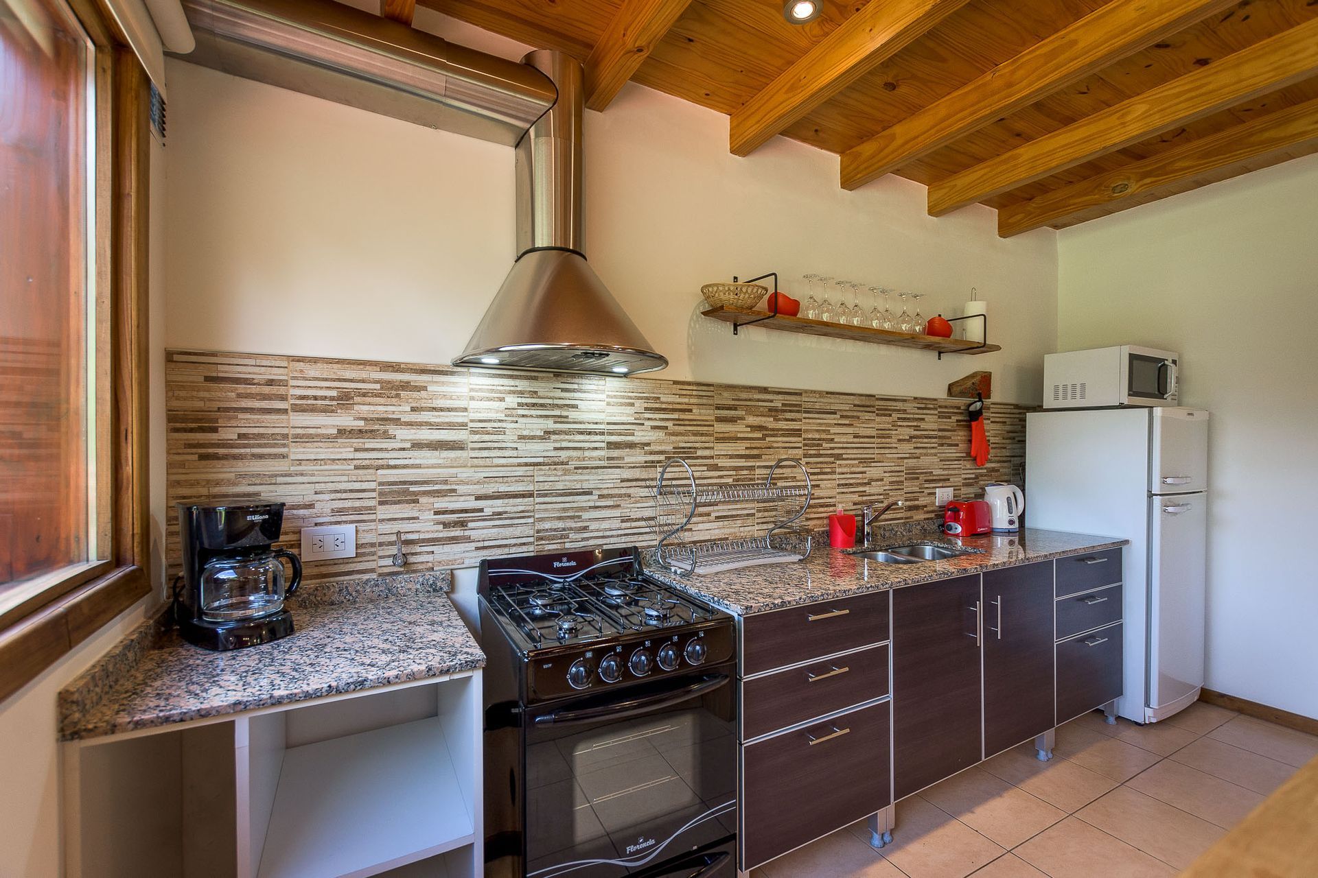 Una cocina rústica con techos con vigas de madera, armarios marrones, cocina de gas, campana extractora de acero inoxidable y nevera blanca.