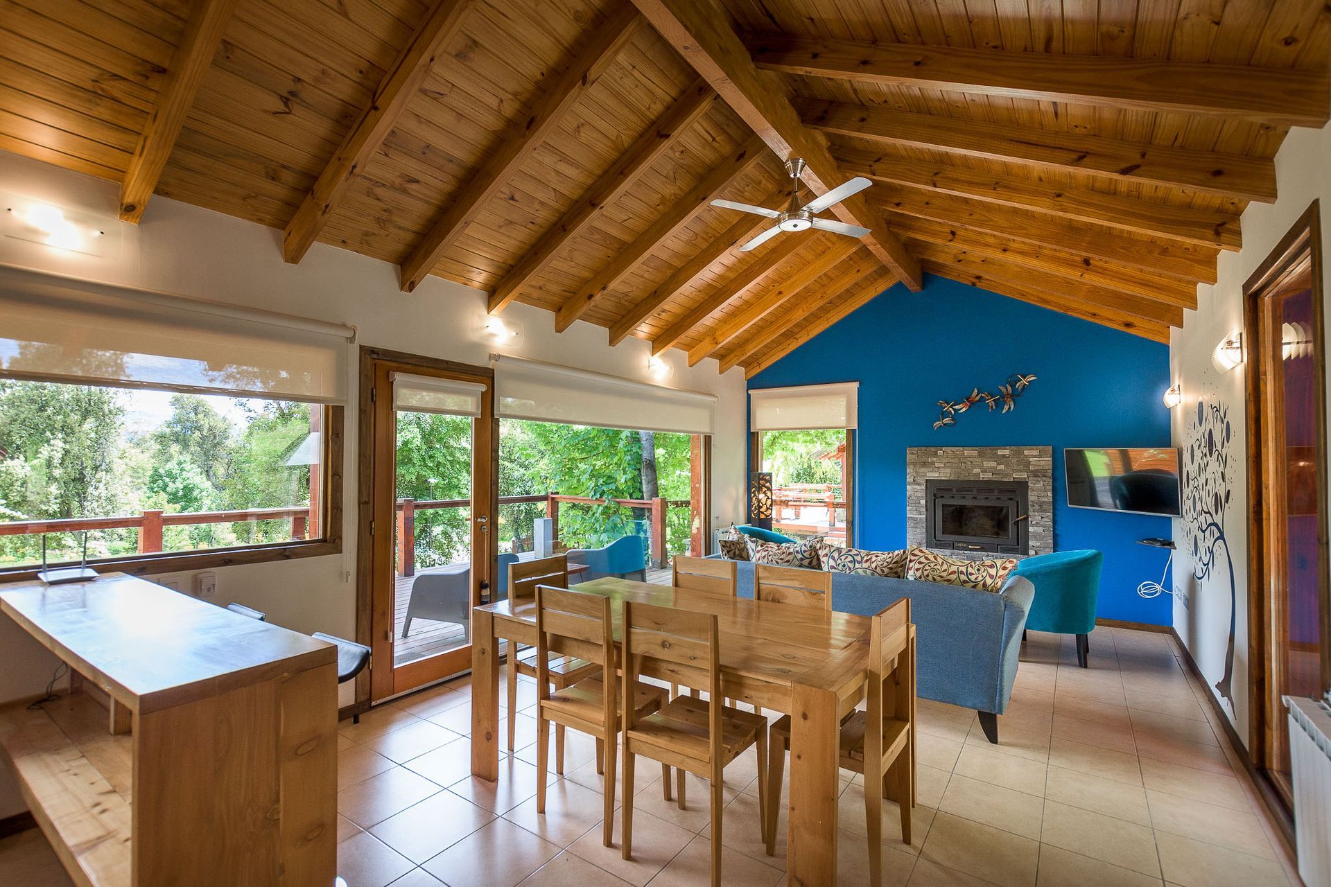 Una luminosa habitación tipo cabaña de planta abierta con techos de madera, zona de comedor, chimenea de piedra y una pared decorativa de color azul.